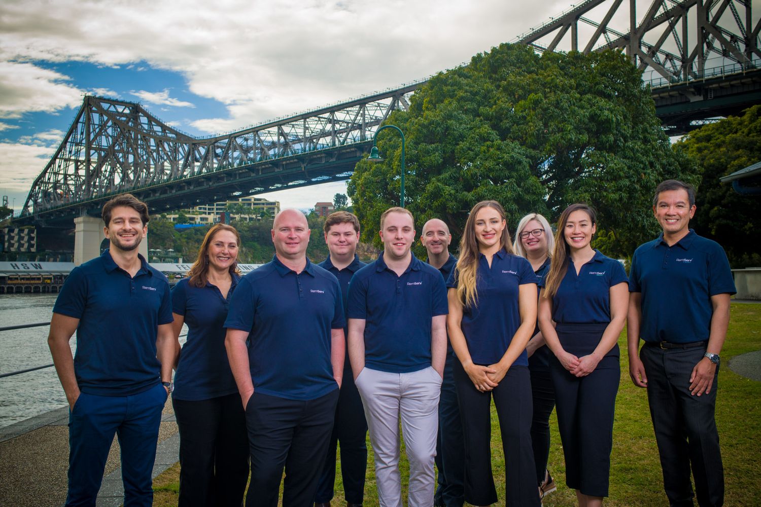 Professional team of ten people in navy polo shirts posing outdoors near iconic Brisbane Story Bridge.