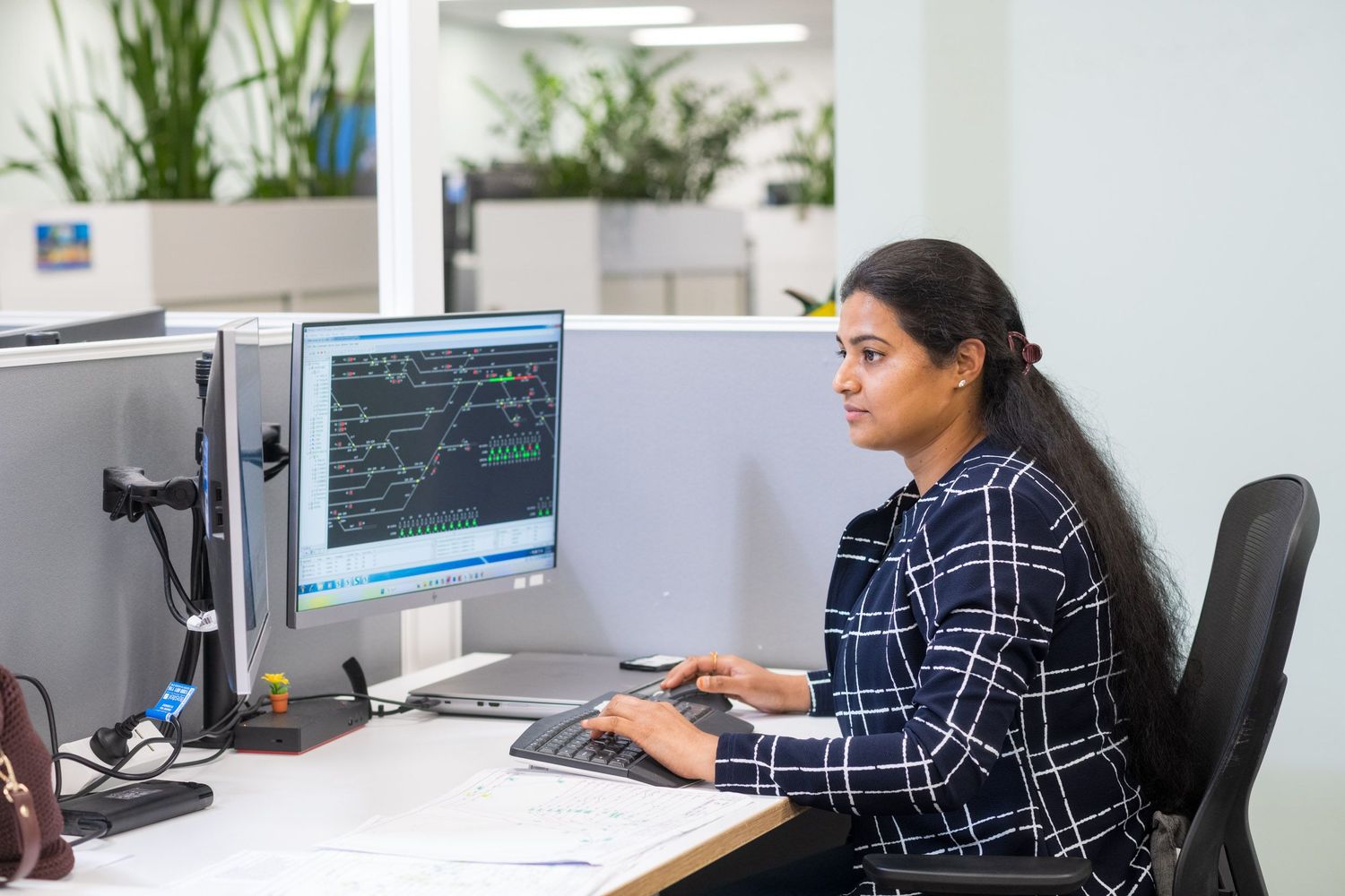 Female professional in checkered shirt works at dual monitor computer workstation in bright office.