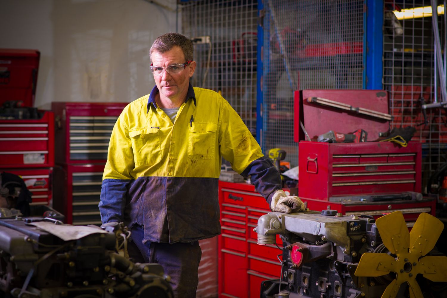 Male technician in yellow hi-vis workwear examining engine parts in a mechanical workshop.