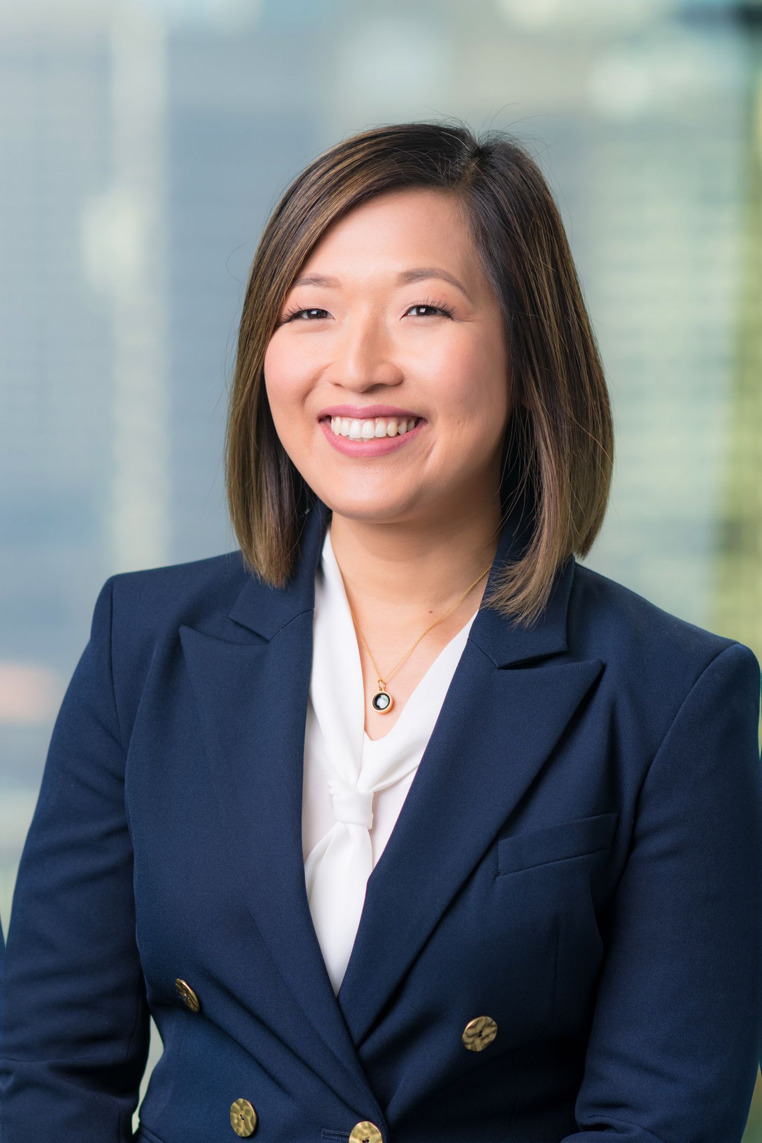 Professional headshot of a smiling Asian woman in a navy blazer with gold buttons and white blouse.