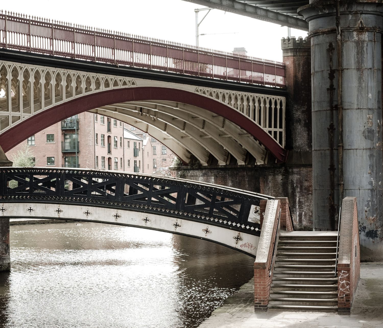Victorian iron bridge with ornate arches spanning urban canal with stone steps and brick pillars.
