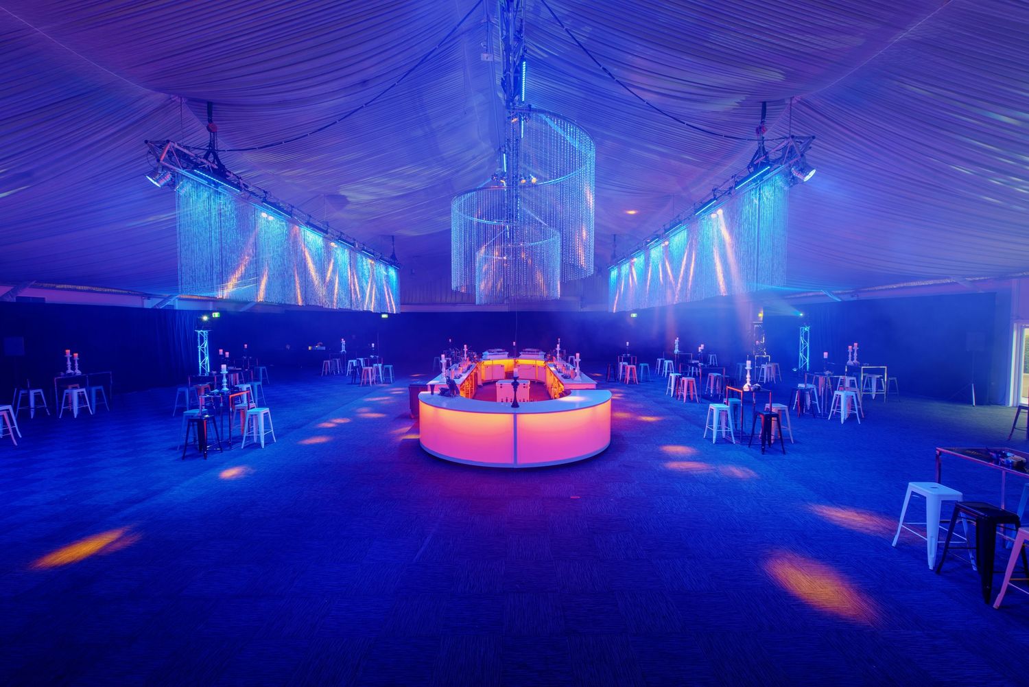 Spacious event tent with blue lighting, a circular illuminated bar, and white high-top tables for a party.