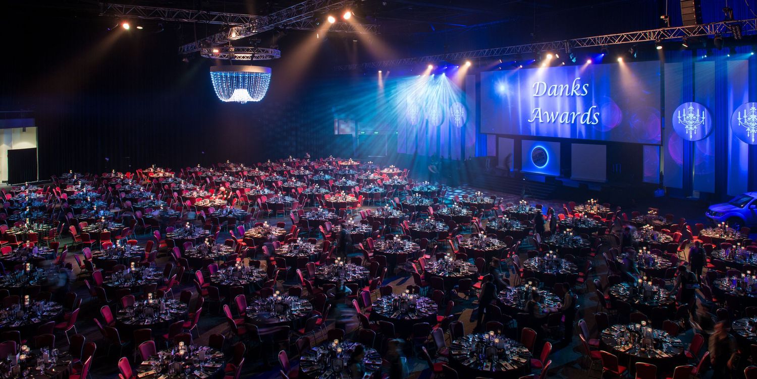 Packed gala dinner venue with round tables and dramatic blue stage lighting for awards night.