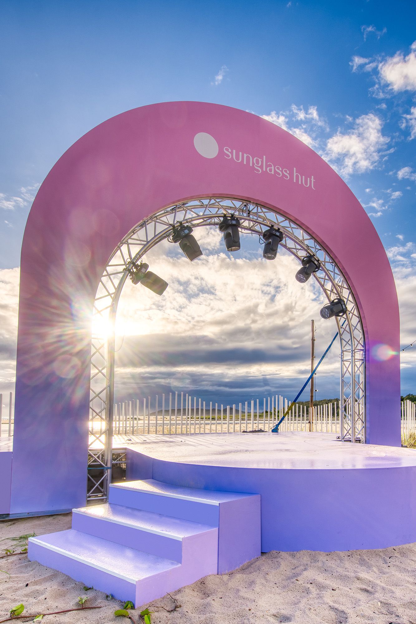 Pink and purple Sunglass Hut branded arch stage on a beach with dramatic sunset sky.