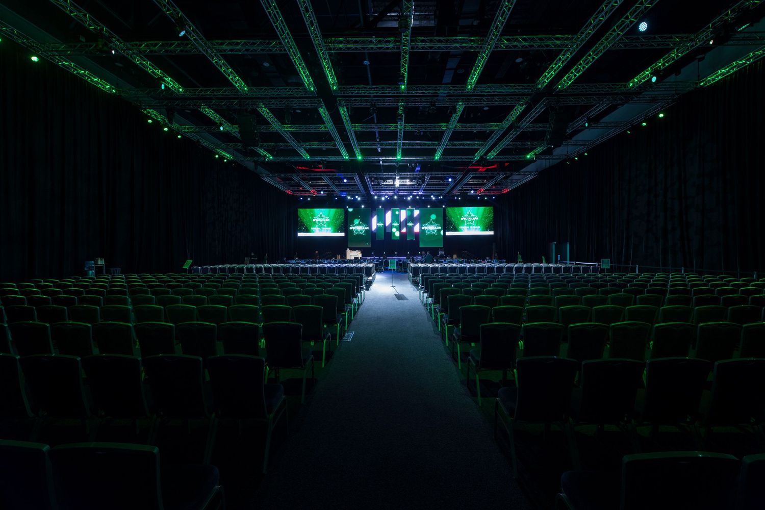 Empty large conference hall with green-lit chairs and dual projection screens.