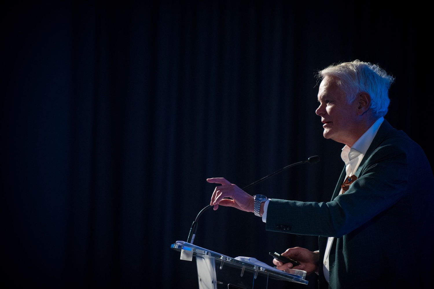 Speaker gesturing at podium during formal conference presentation on dark stage.
