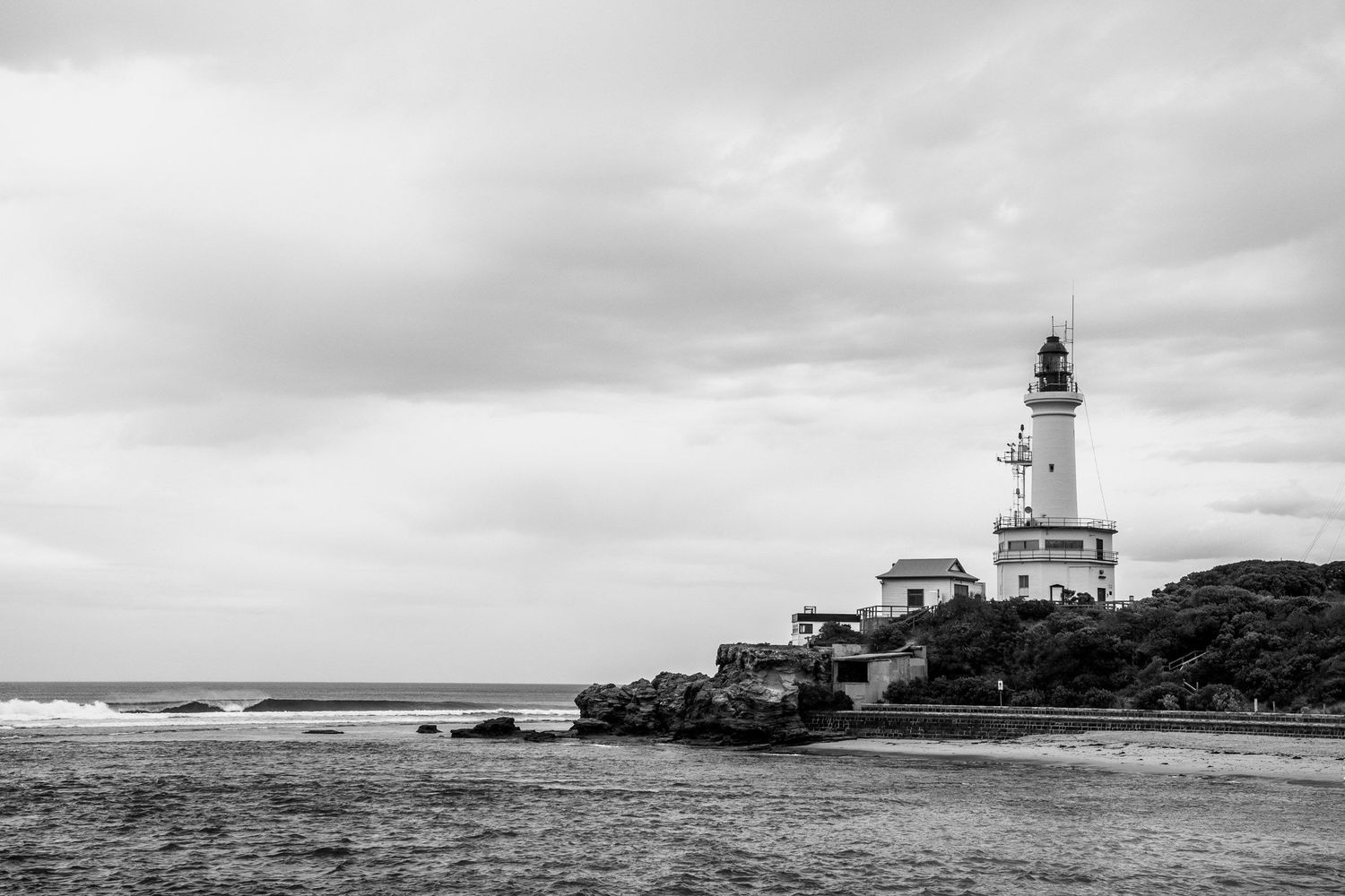 Black and white coastal landscape featuring a historic white lighthouse on rocky shoreline under cloudy skies.