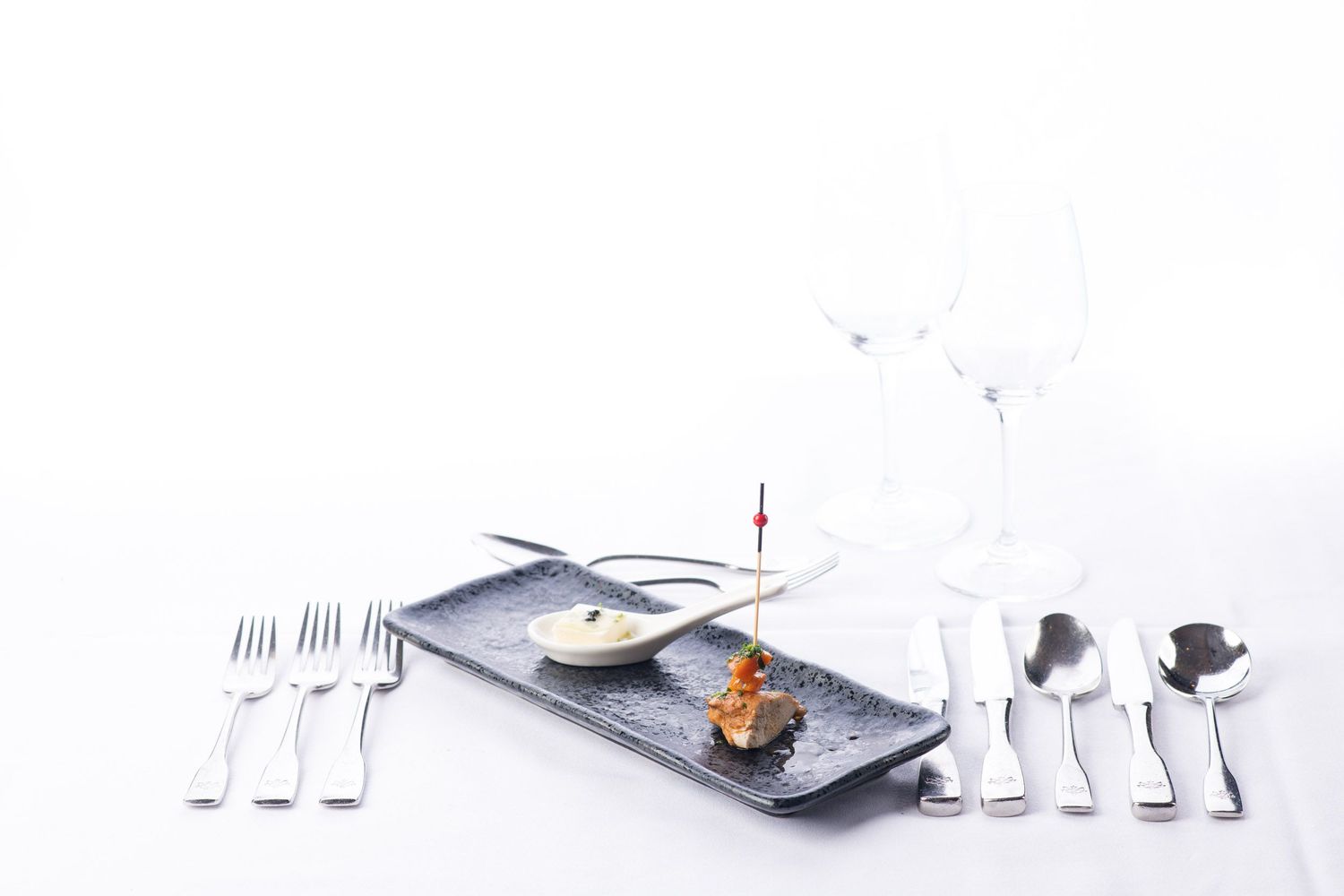 Elegant restaurant place setting with small gourmet appetizer on black rectangular plate with crystal glasses.
