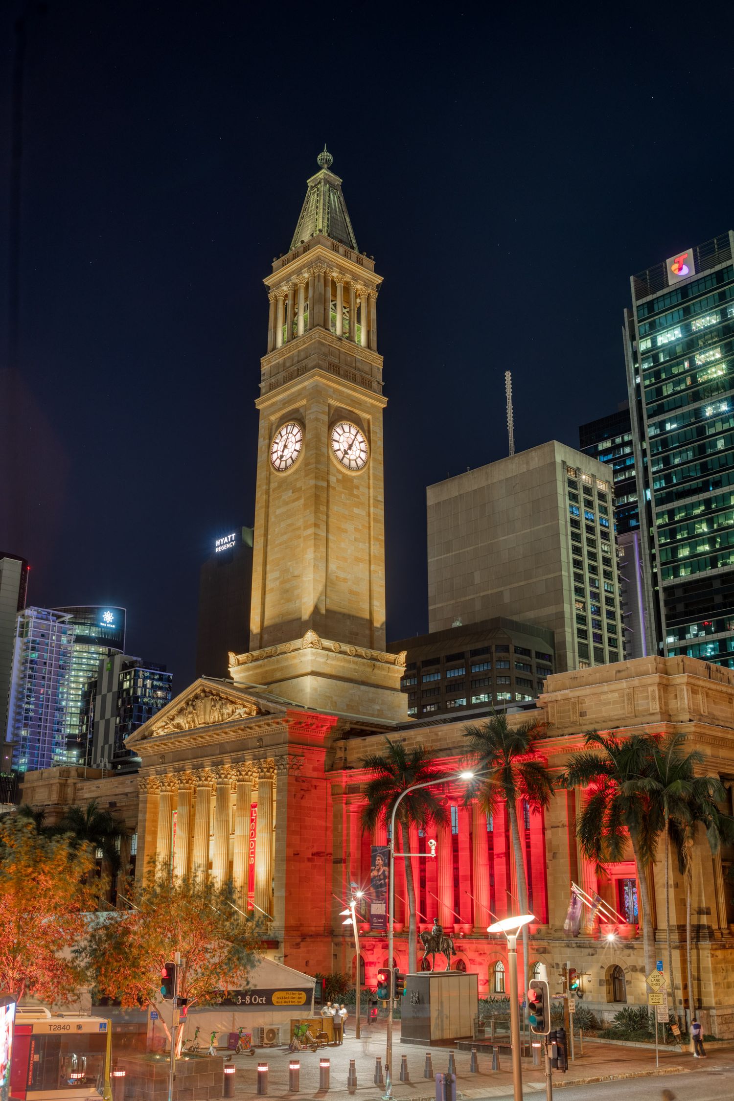 Brisbane City Hall clock tower lit in warm gold and red lights at night surrounded by modern CBD skyscrapers.