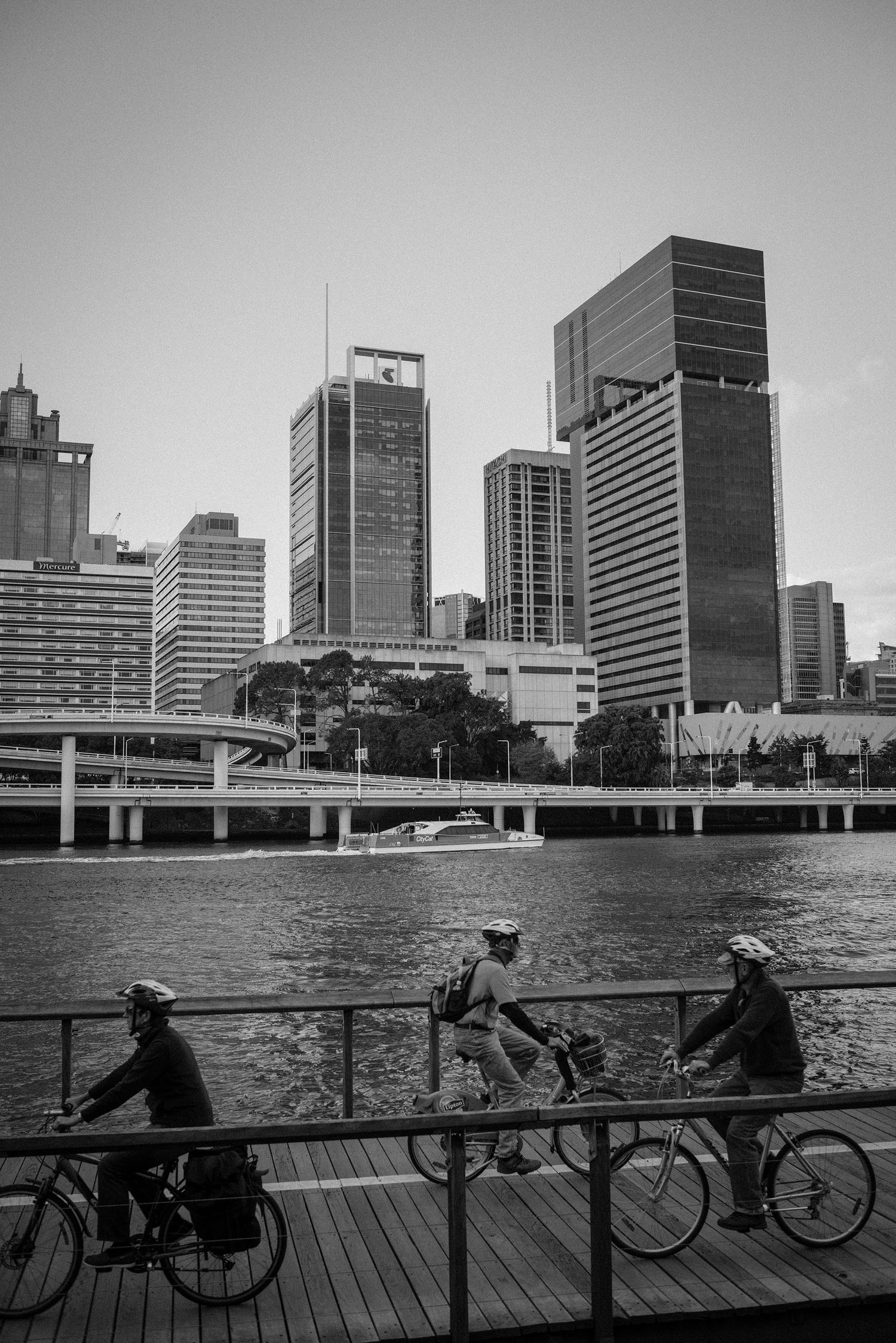 Black and white photo of three cyclists riding along a riverside boardwalk with city skyline behind.