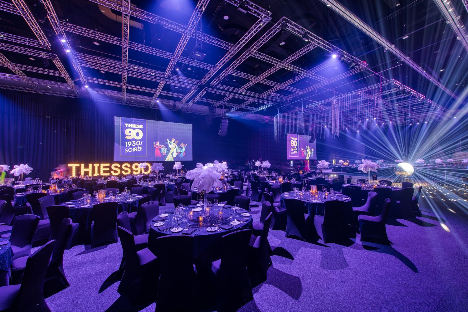 Thiess 90th anniversary gala dinner with purple lighting, round tables, and illuminated signage in a large event venue.