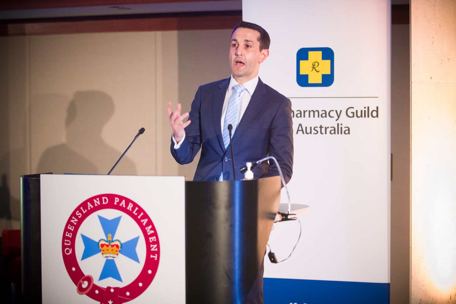 Male speaker presenting at a podium with Queensland Parliament and Pharmacy Guild of Australia branding.