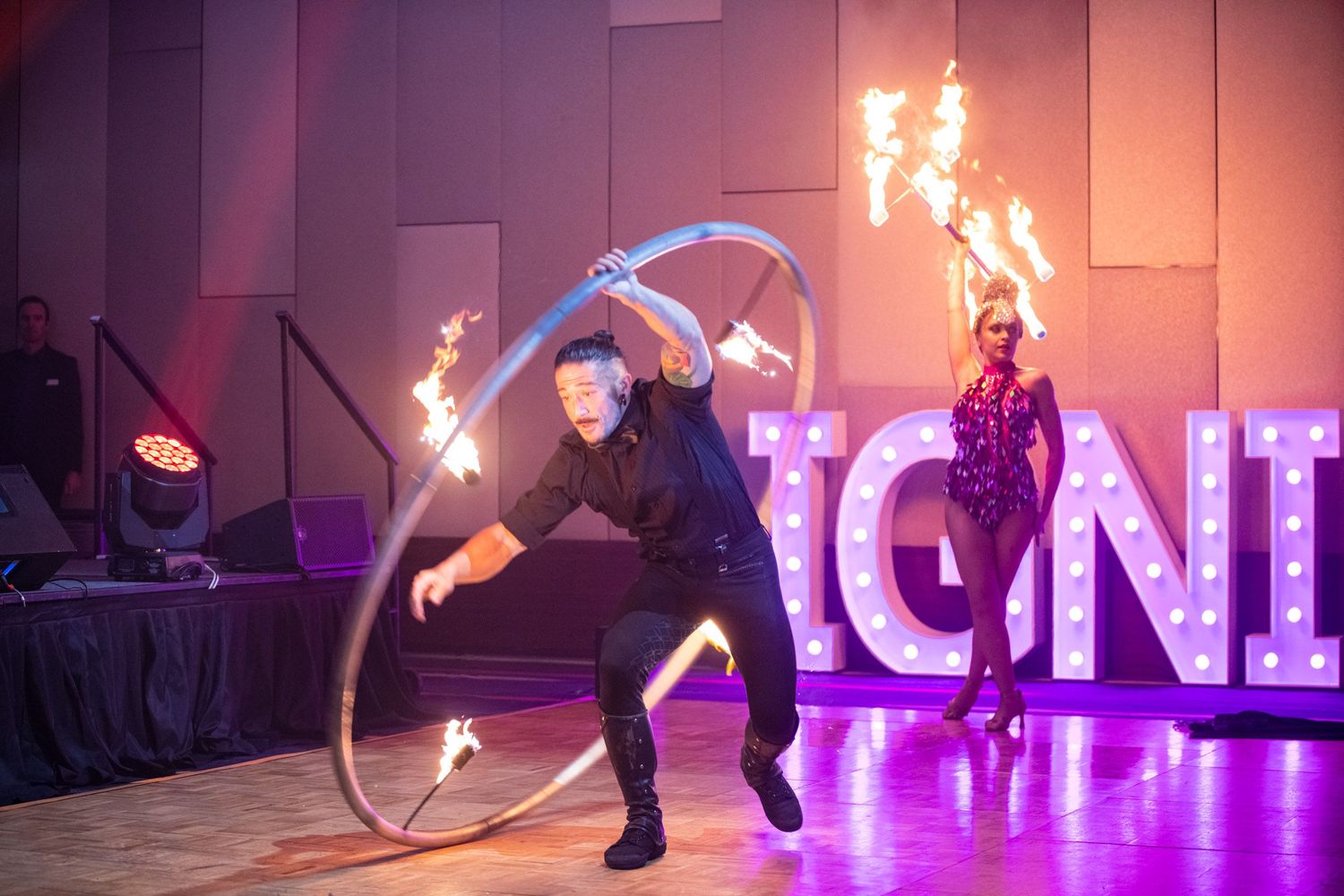 Male performer spinning fire hoop on stage with female fire dancer beside illuminated IGNITE sign at gala.