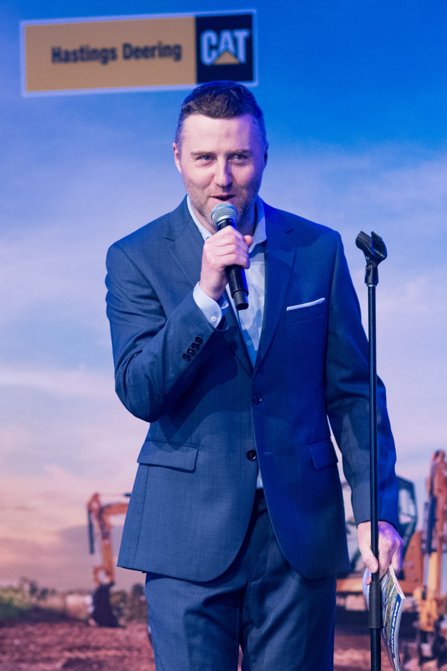 Professional male MC in blue suit speaking into microphone at Hastings Deering CAT corporate event.
