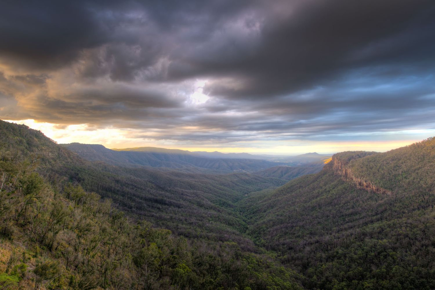 Dramatic sunset over vast forested mountain valley with stormy clouds and golden light breaking through.
