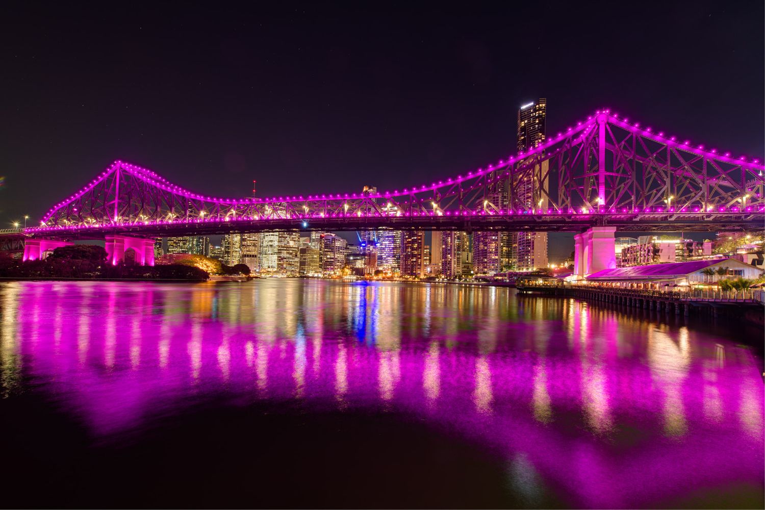 Story Bridge in Brisbane illuminated in vibrant pink and purple lights reflecting on the river at night.