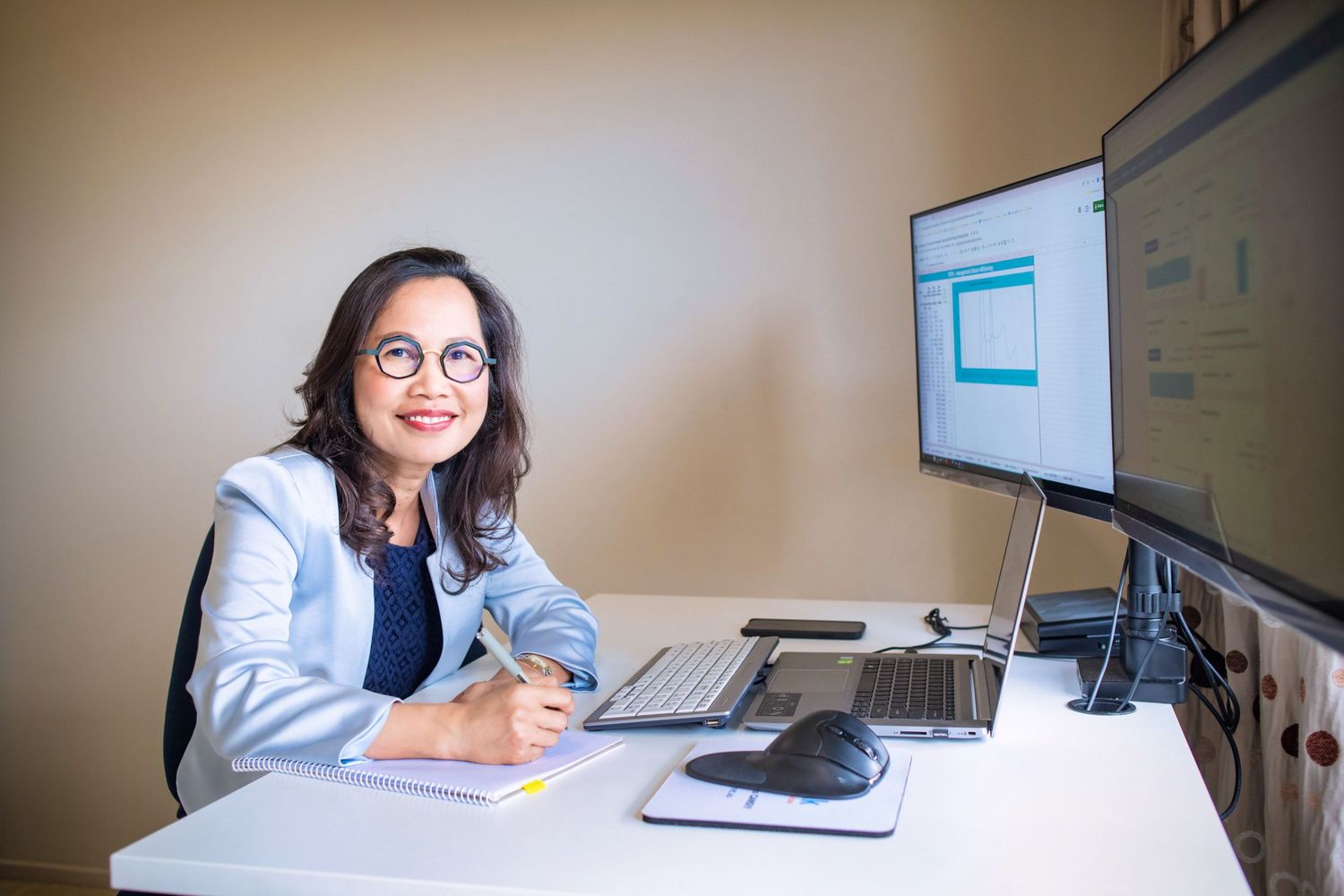 Professional woman in blue blazer smiling at desk with dual monitors in a modern office.