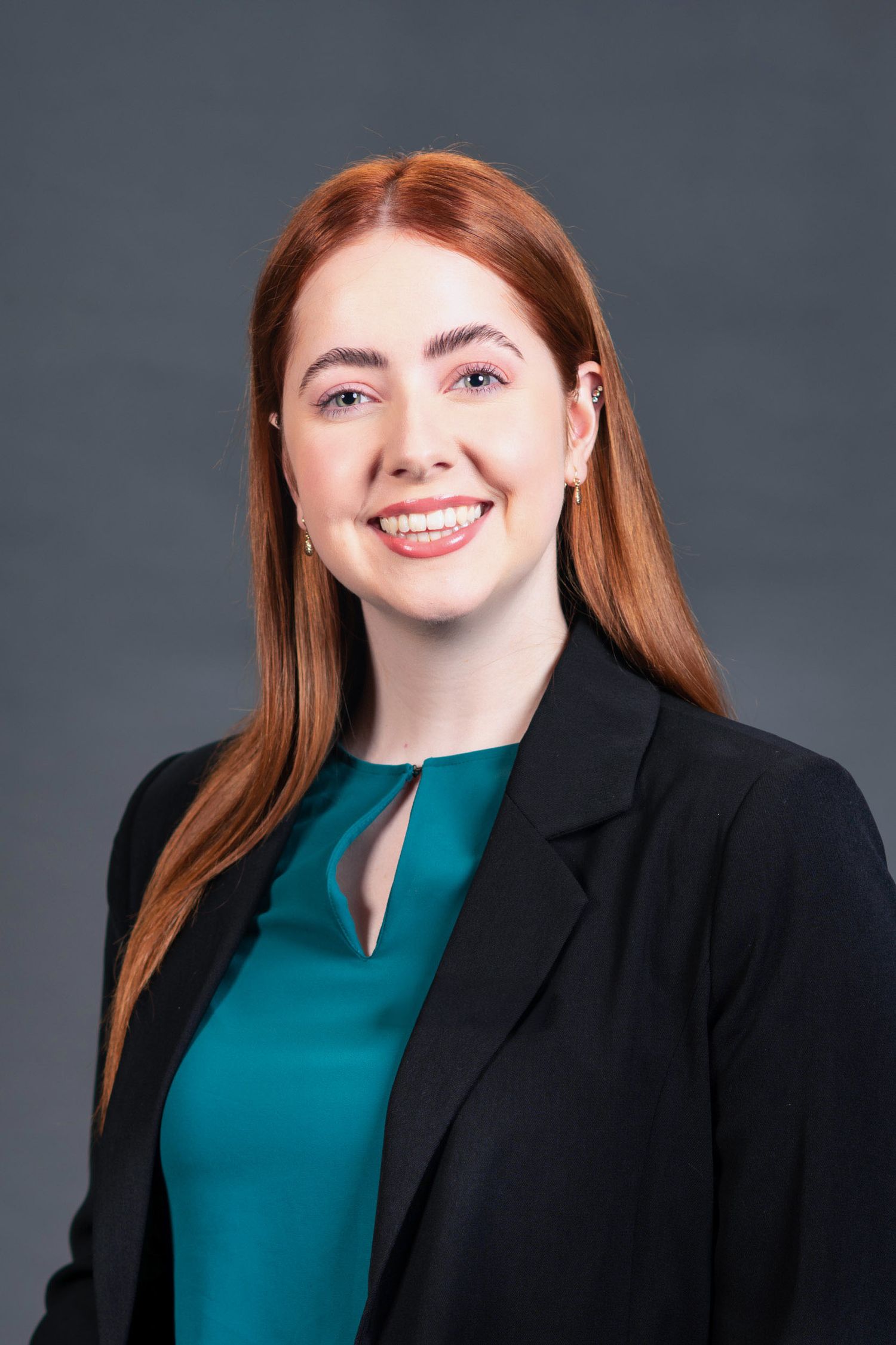 Smiling young woman with long red hair wearing a teal shirt and black blazer on gray background.