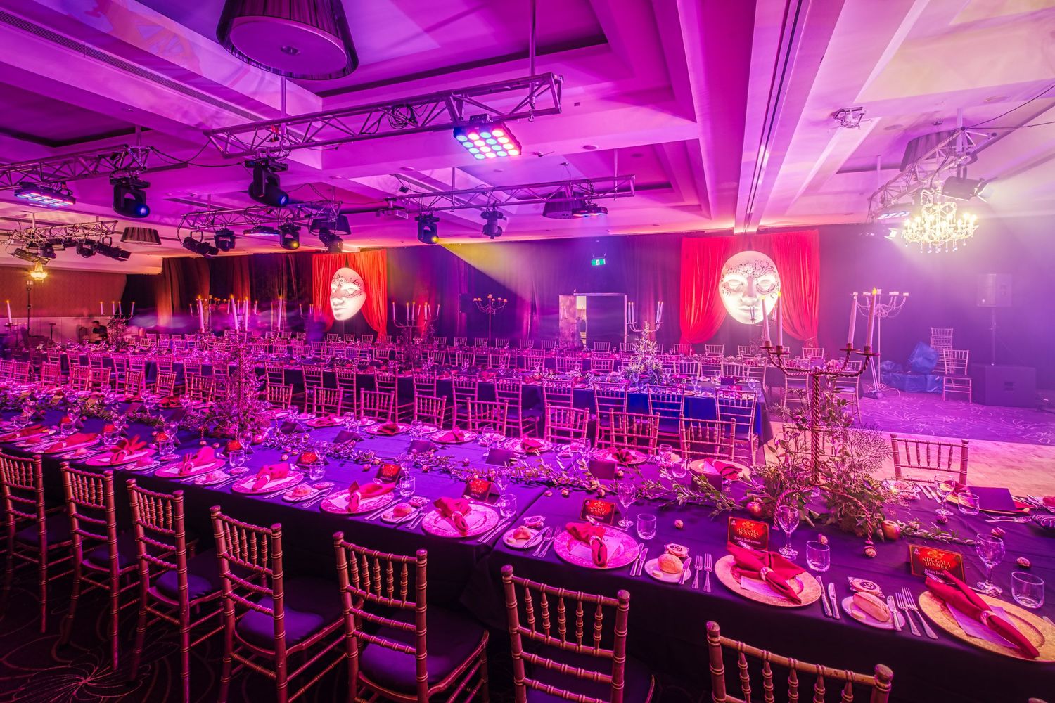 Luxurious gala dinner venue with long decorated tables, gold chiavari chairs, and dramatic pink and purple lighting.