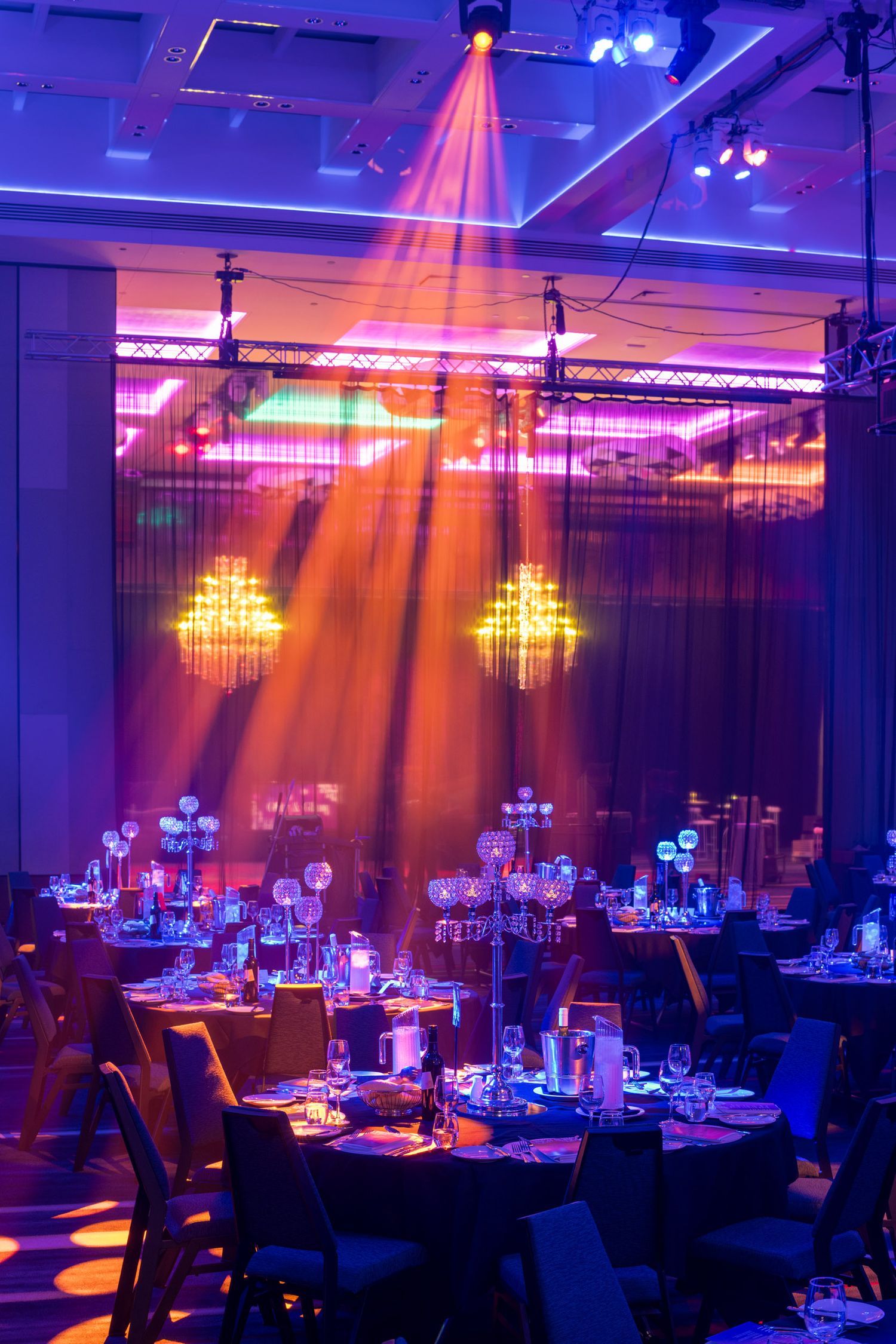 Elegant gala dinner setup with purple and orange lighting, chandeliers, and round tables in a grand ballroom.