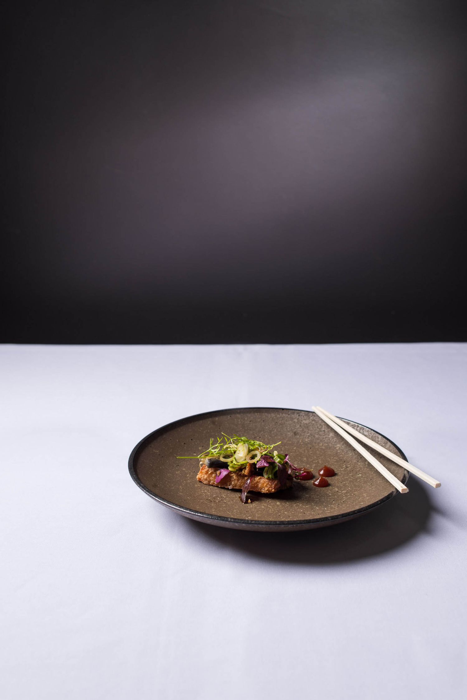 Japanese-inspired glazed meat dish garnished with green onions and chopsticks on a dark ceramic plate.