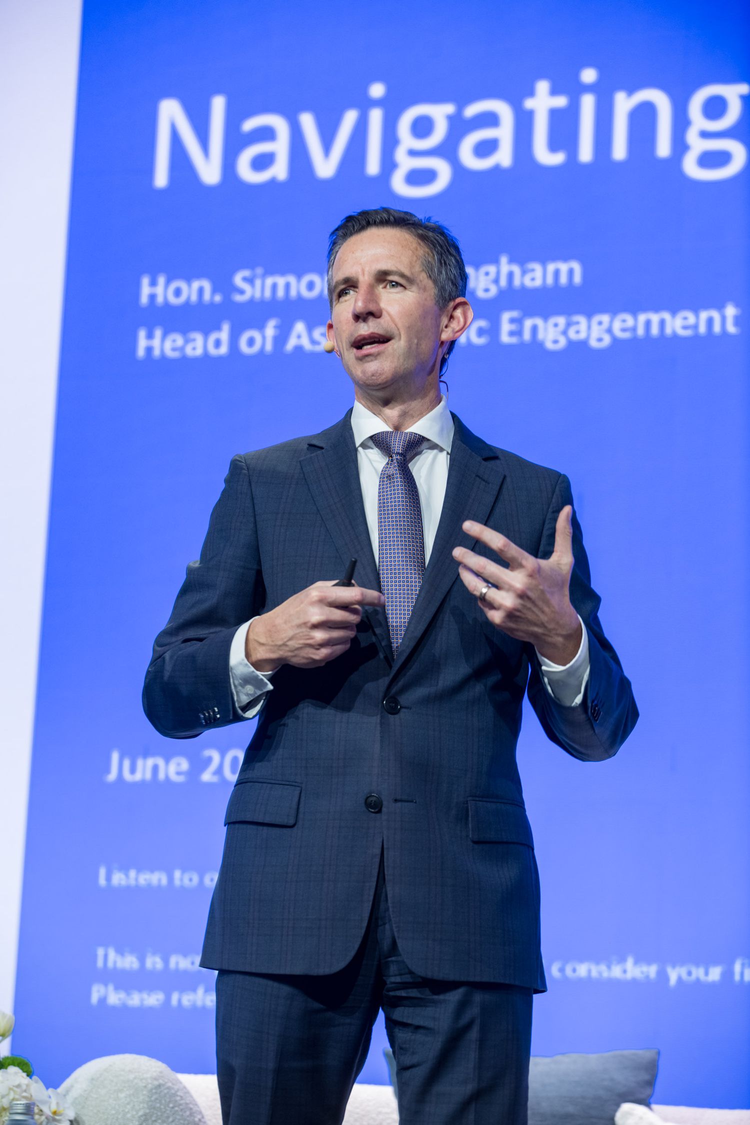 Suited male speaker presents at podium before blue Navigating conference backdrop screen.