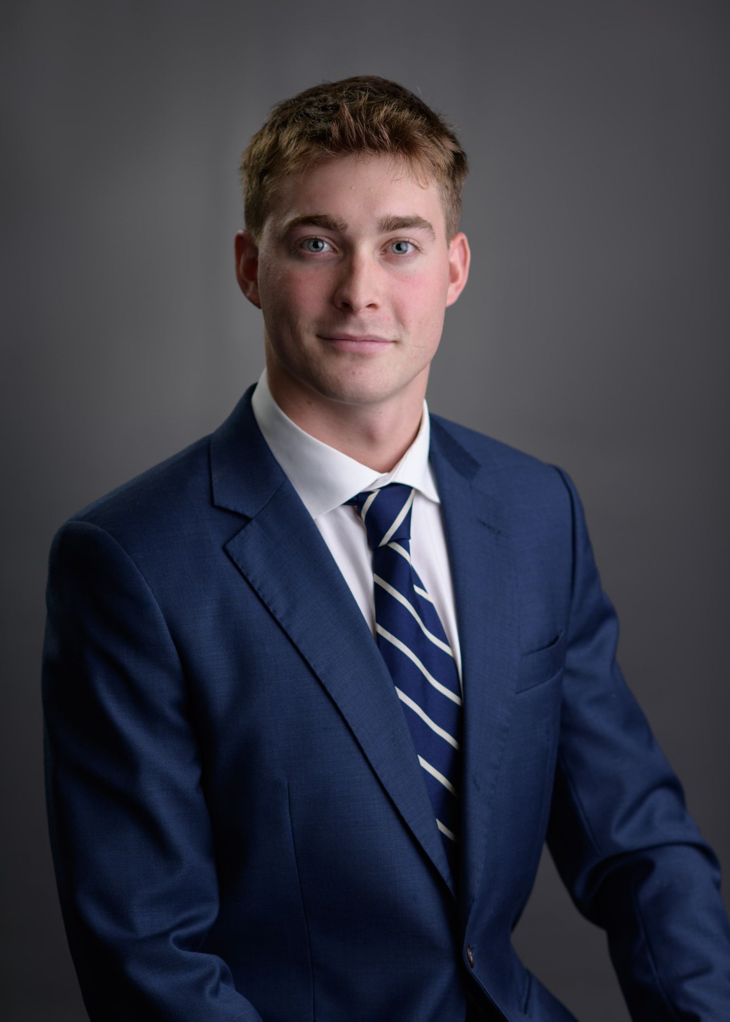 Professional headshot of a young man in a navy blue suit with a striped tie.