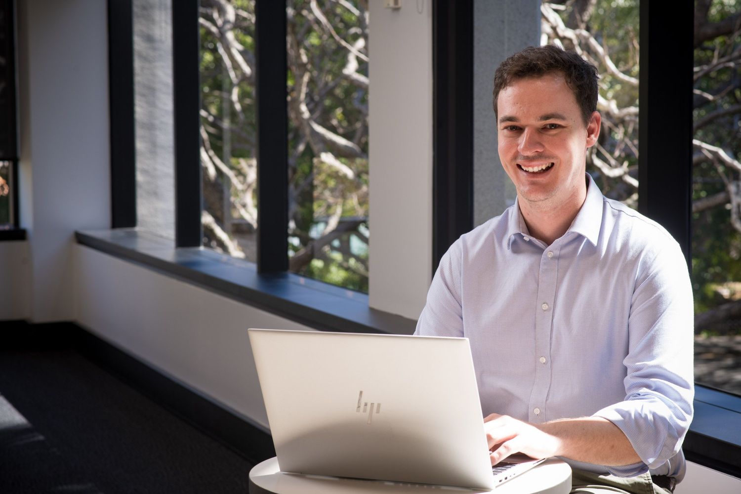 Young professional man smiles while working on HP laptop in modern office with large windows.