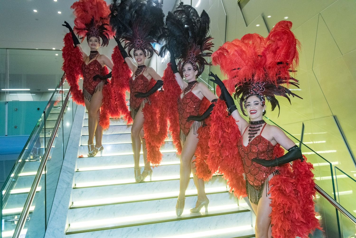 Showgirls in vibrant red feathered costumes and headdresses posing dramatically on a staircase at a gala event.