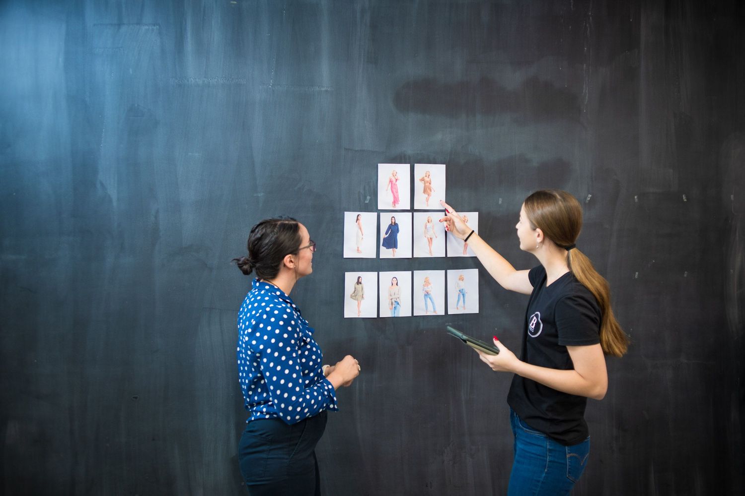 Two women reviewing fashion design sketches pinned to a dark wall in a creative studio setting.
