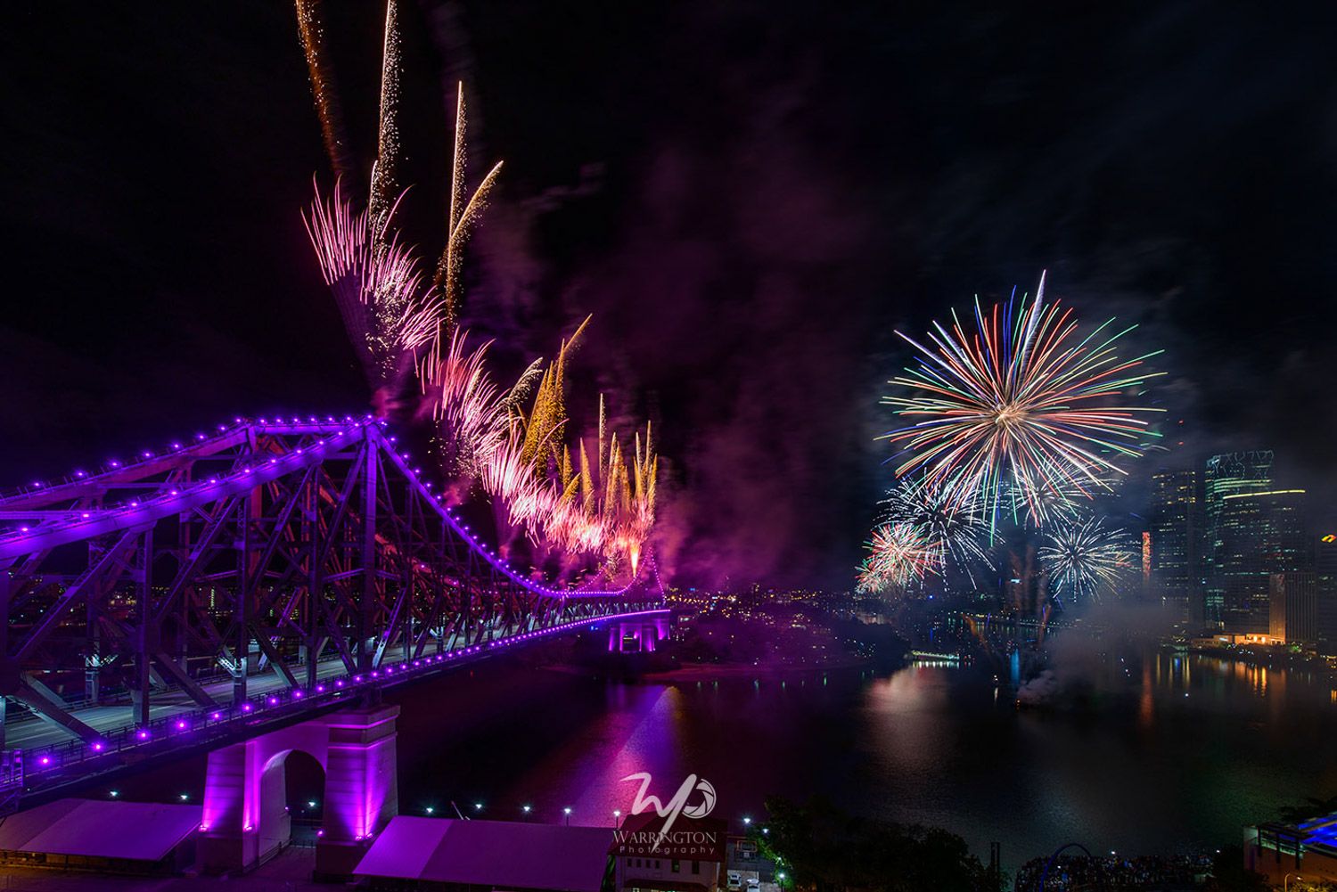 Colourful fireworks display over illuminated Story Bridge Brisbane reflecting on river at night.