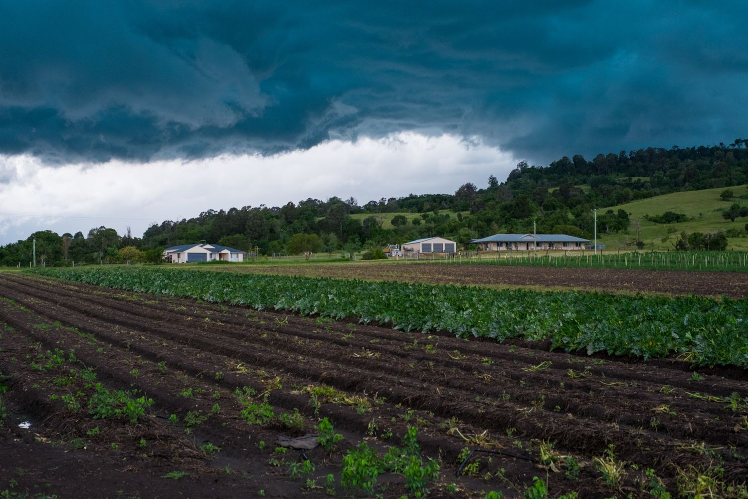 Dark storm clouds loom over freshly plowed farm fields with rural buildings and green hills.