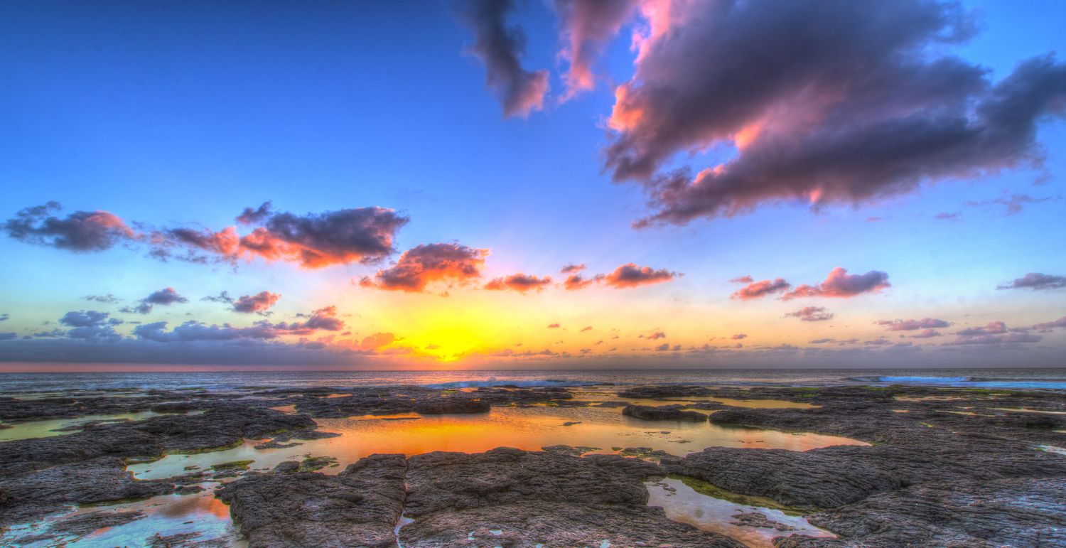Panoramic HDR sunset over rocky shore with vivid blue sky, dramatic clouds, and golden sun reflecting on water.