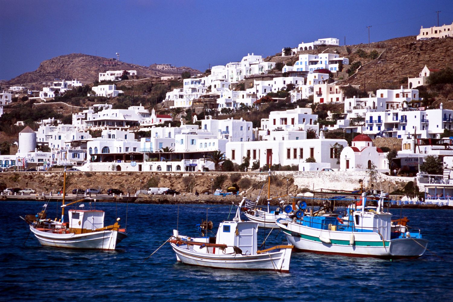 Traditional fishing boats moored in harbor with white Cycladic buildings on Mykonos, Greece.