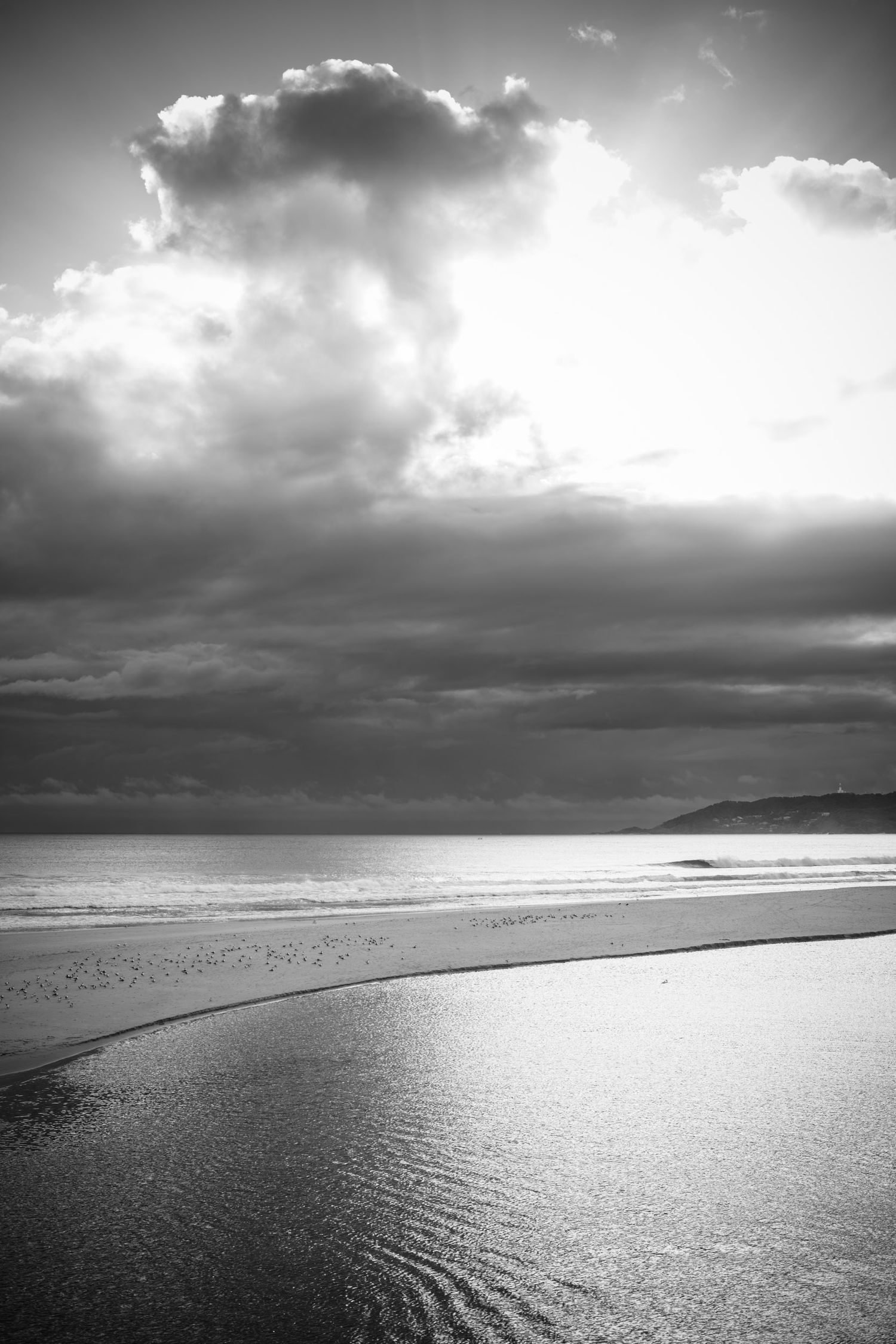 Black and white dramatic coastal beach scene with moody storm clouds and sunlight breaking through.
