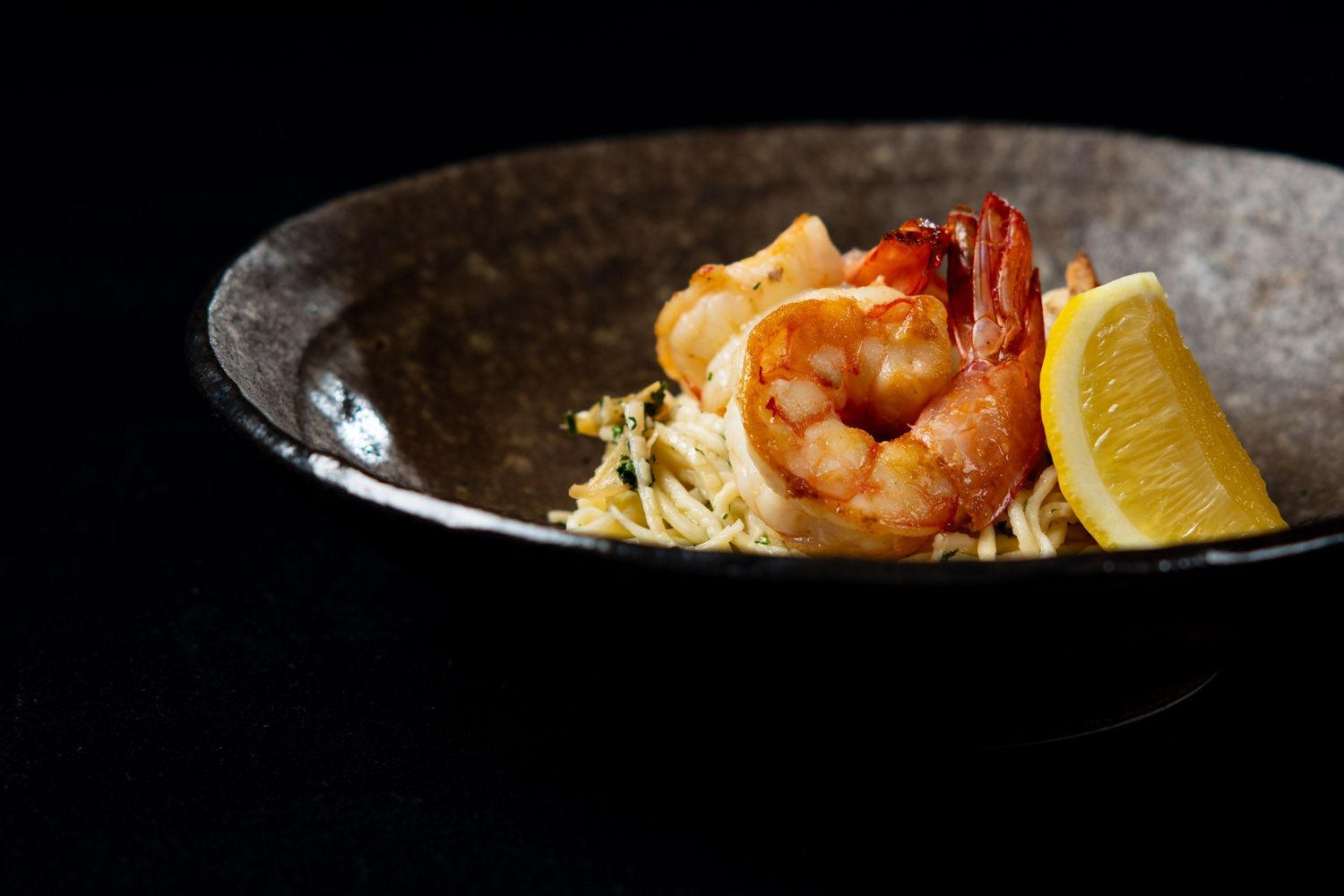 Grilled shrimp with pasta and lemon wedge served in a rustic dark ceramic bowl.