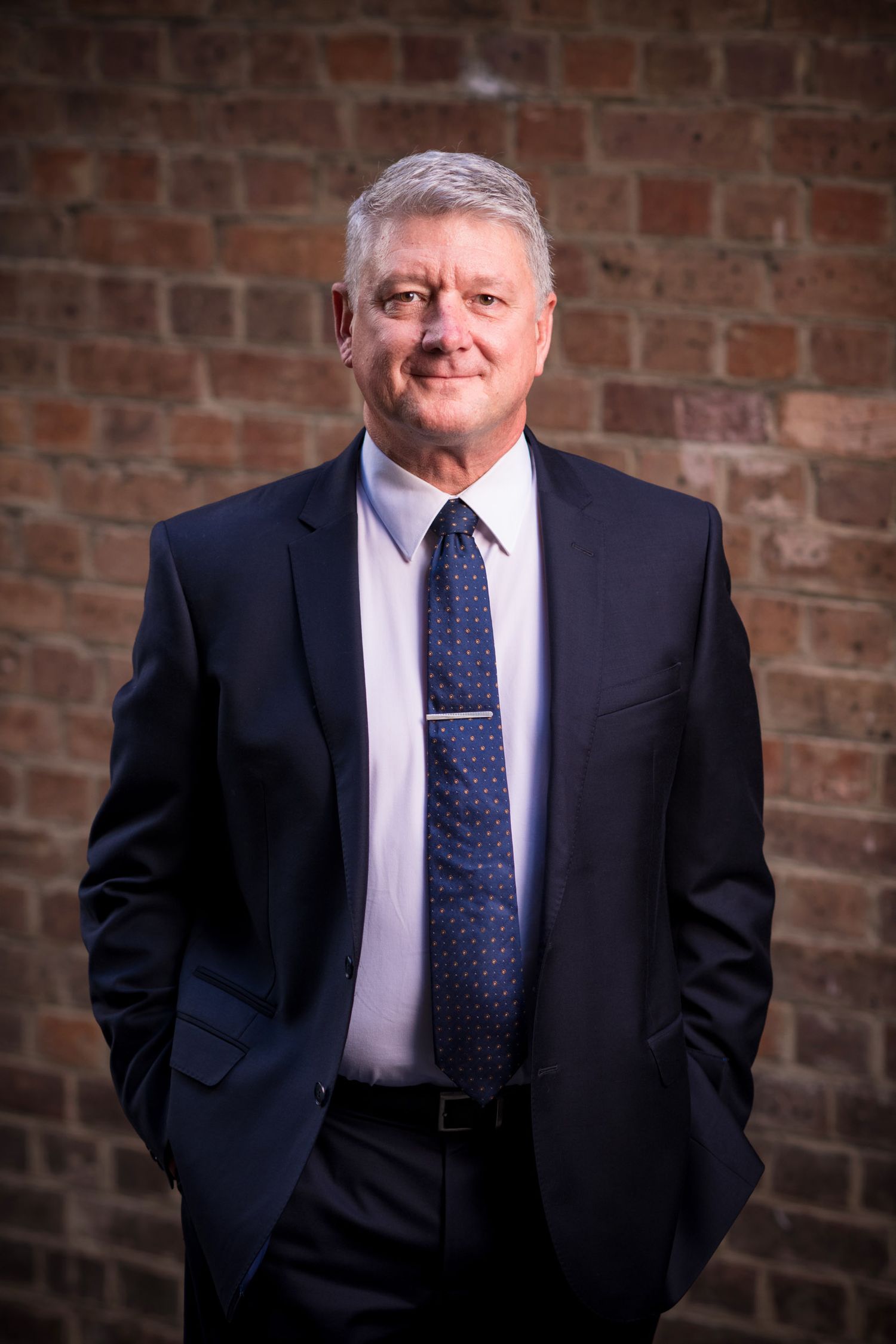 Smiling senior businessman in navy suit and tie, professional portrait against brick wall background.