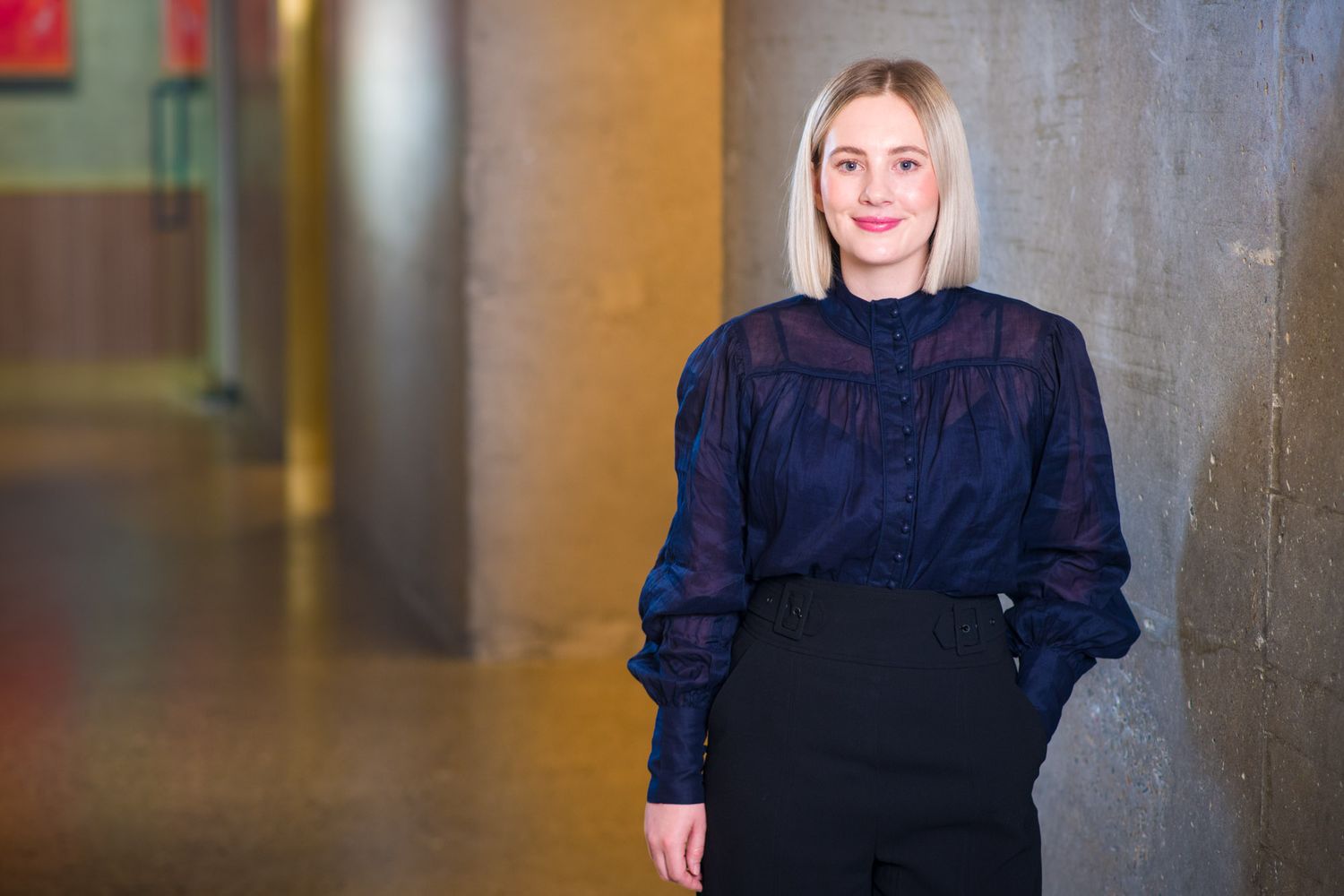 Blonde woman in navy blouse and black trousers leans against concrete wall in hallway.
