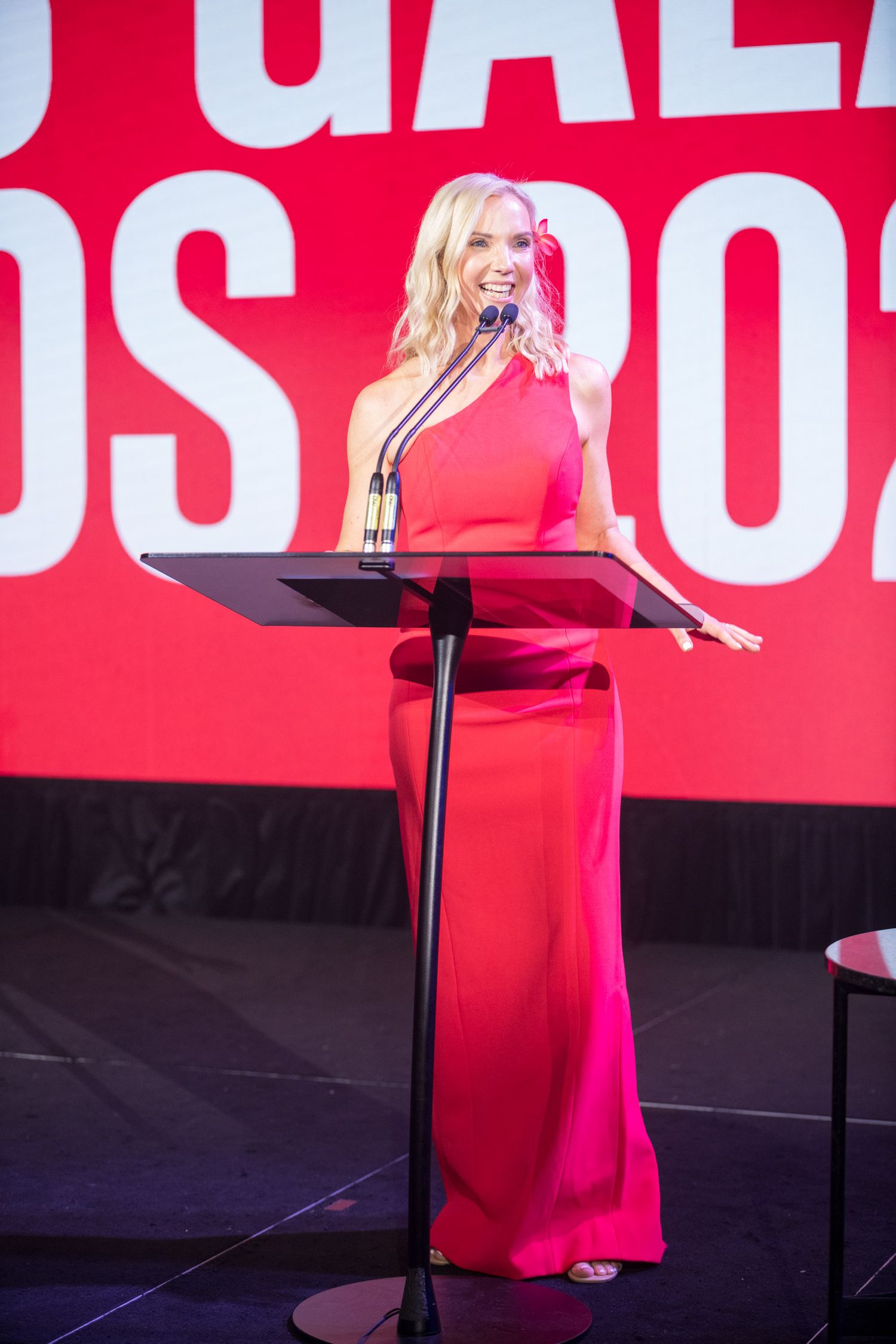 Blonde woman in a red halter gown speaking at a podium during a gala awards ceremony.