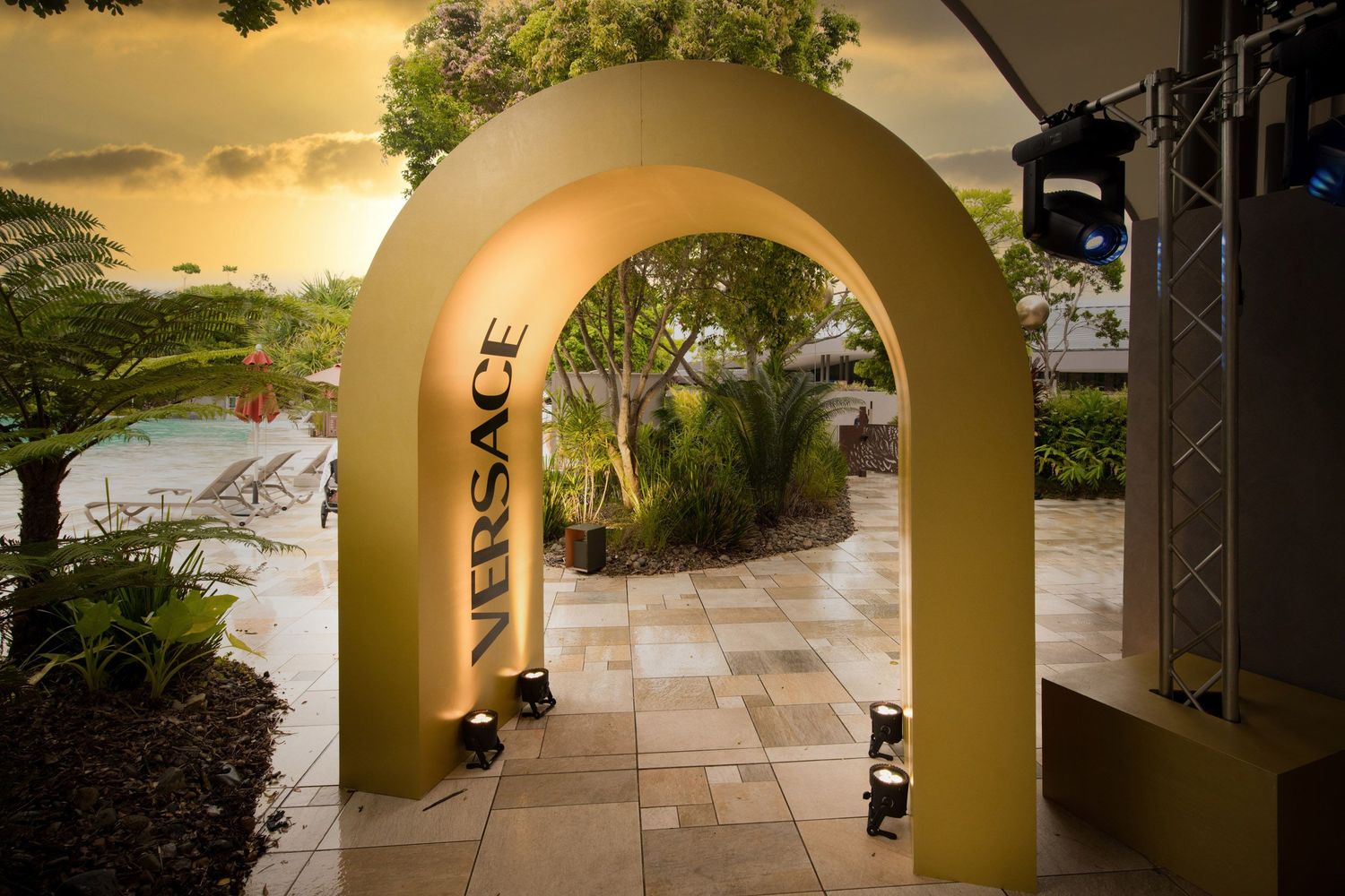 Gold Versace branded arch entrance with tropical garden and pool at sunset.