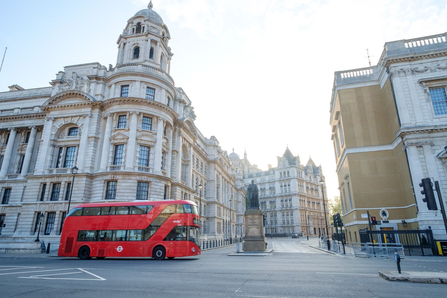 A red double-decker bus passes historic neoclassical buildings on a quiet London street in morning light.