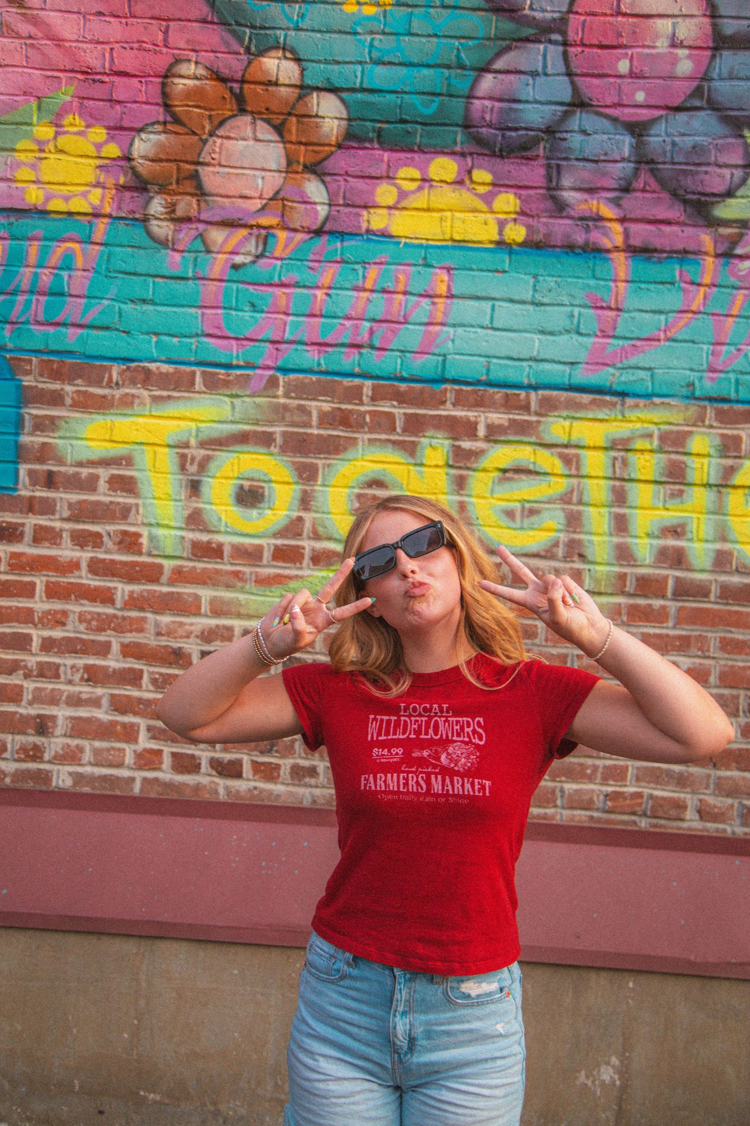 Blonde teen in red Farmers Market tee flashes peace signs wearing sunglasses before colorful mural.