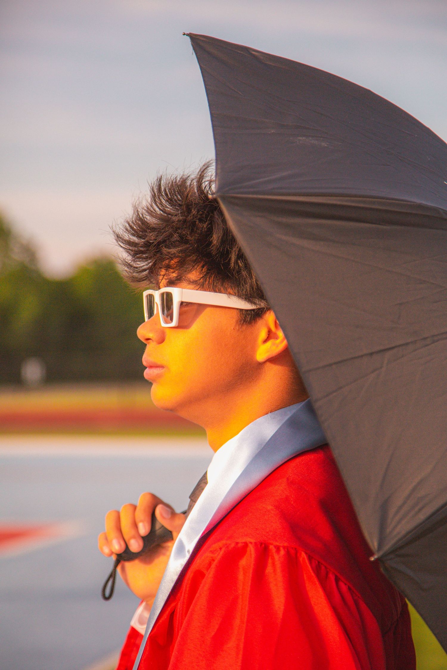 Graduate in red gown holds black umbrella wearing white sunglasses in golden hour light.