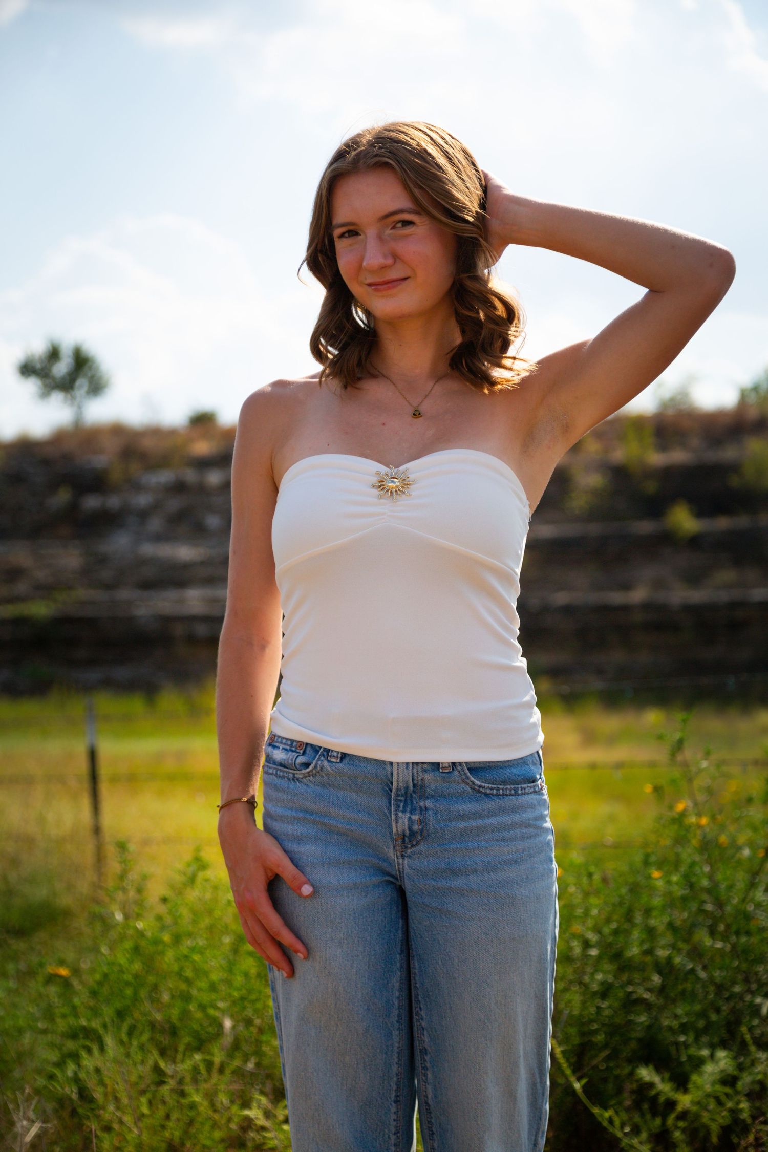 Young woman in white tube top and jeans posing outdoors in a sunny green field with rocky backdrop.