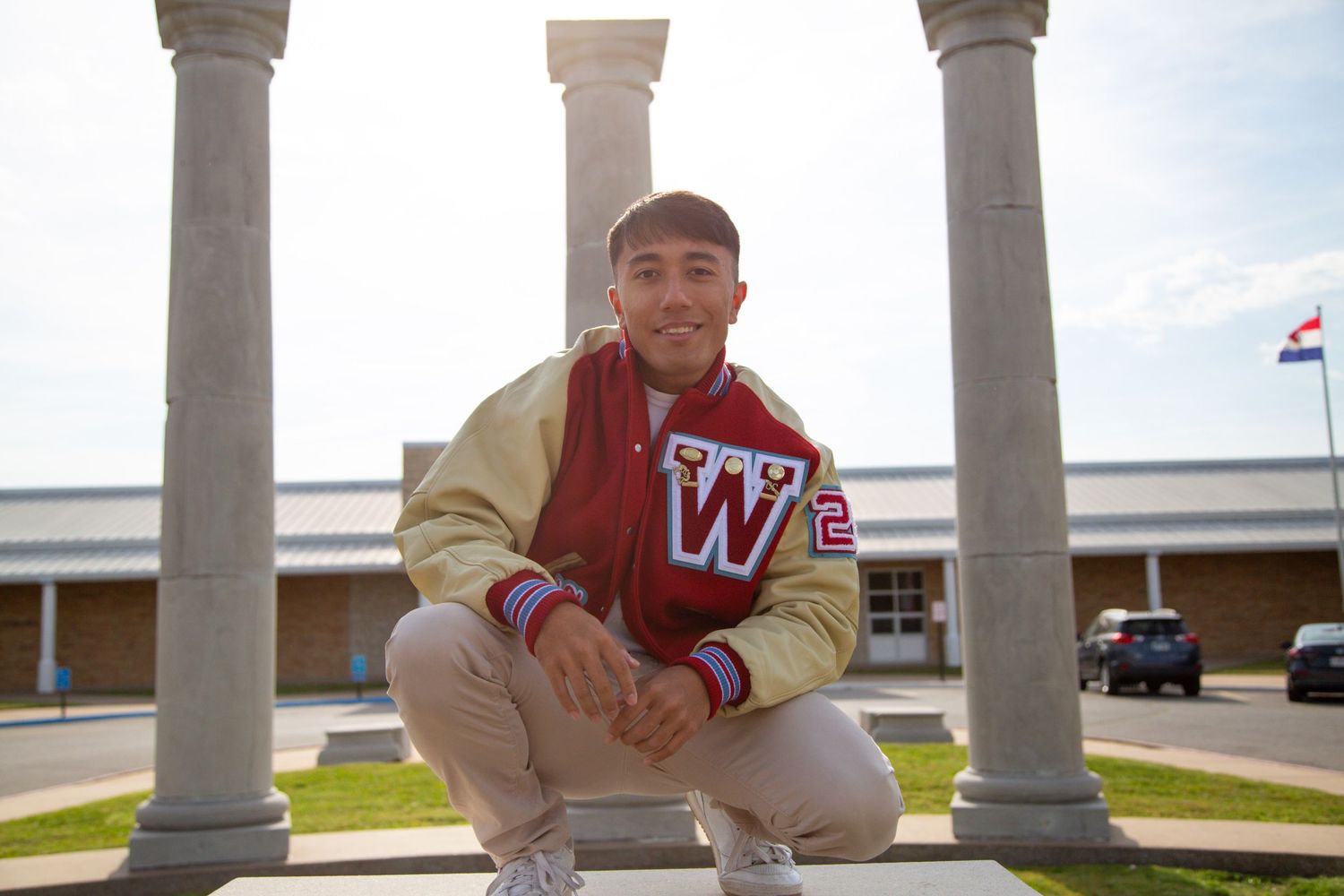 A young man in a red and gold varsity jacket crouches between columns outside Webb City High School.