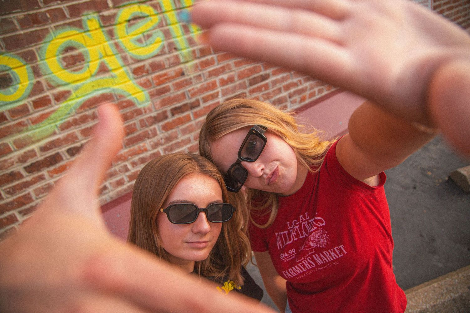 Two young women with sunglasses frame the camera with their hands against a colorful graffiti wall.