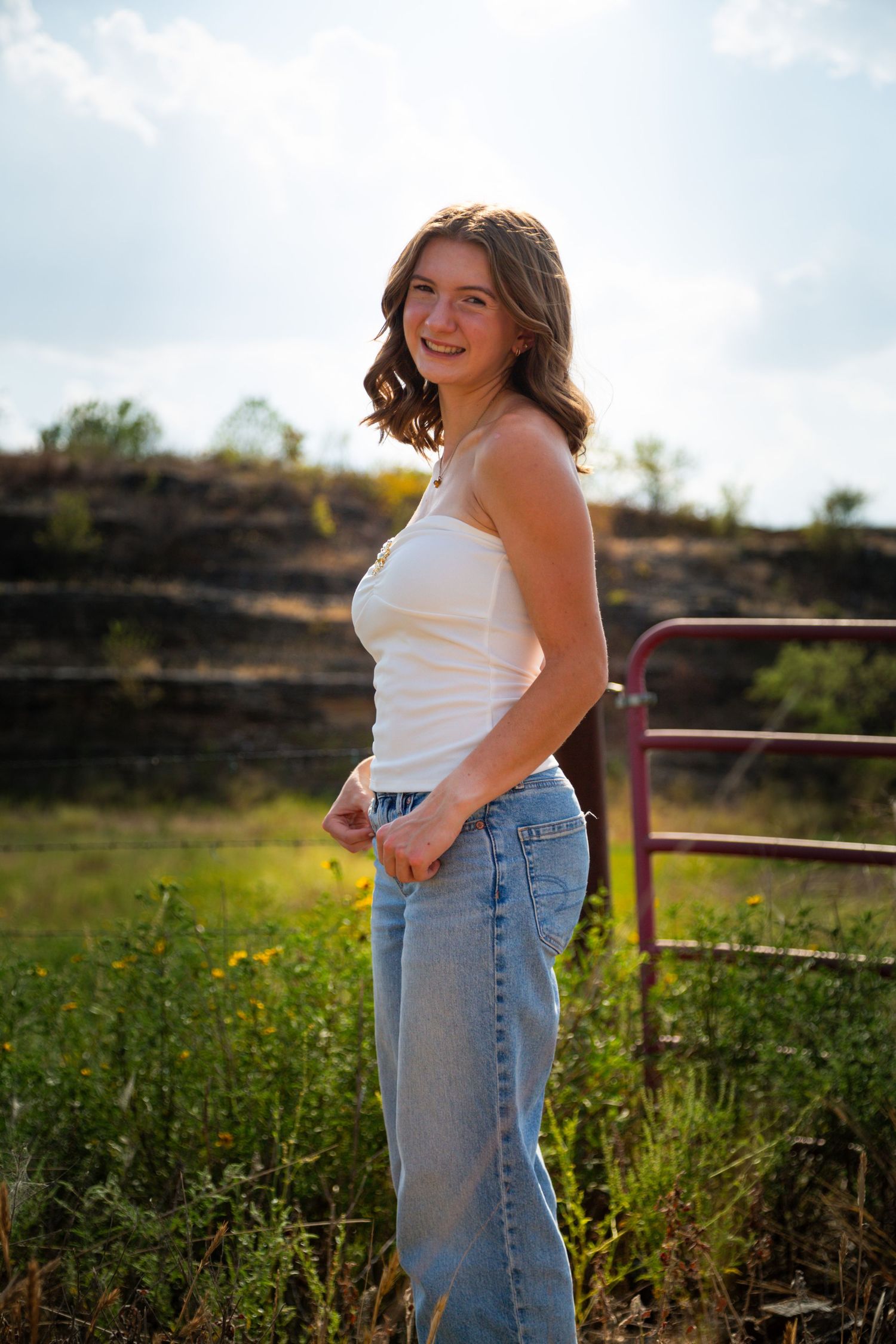 Young woman in white tank top and blue jeans smiling outdoors near a red gate with green fields behind her.