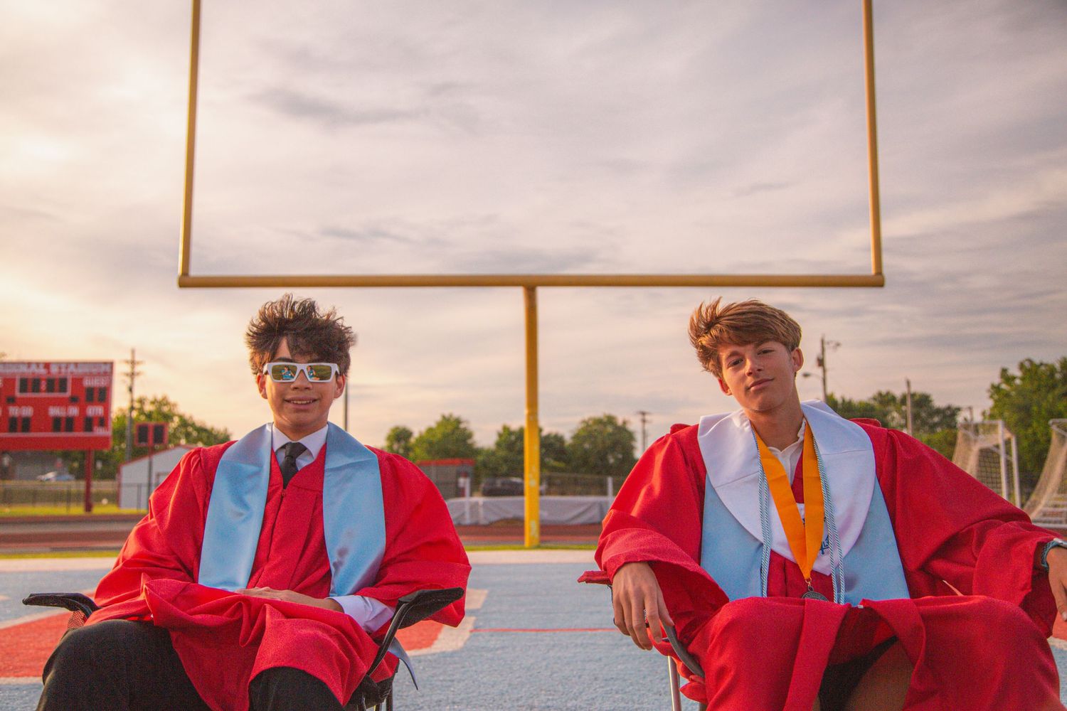 Two male graduates in red gowns sit on football field with goal post at golden hour.