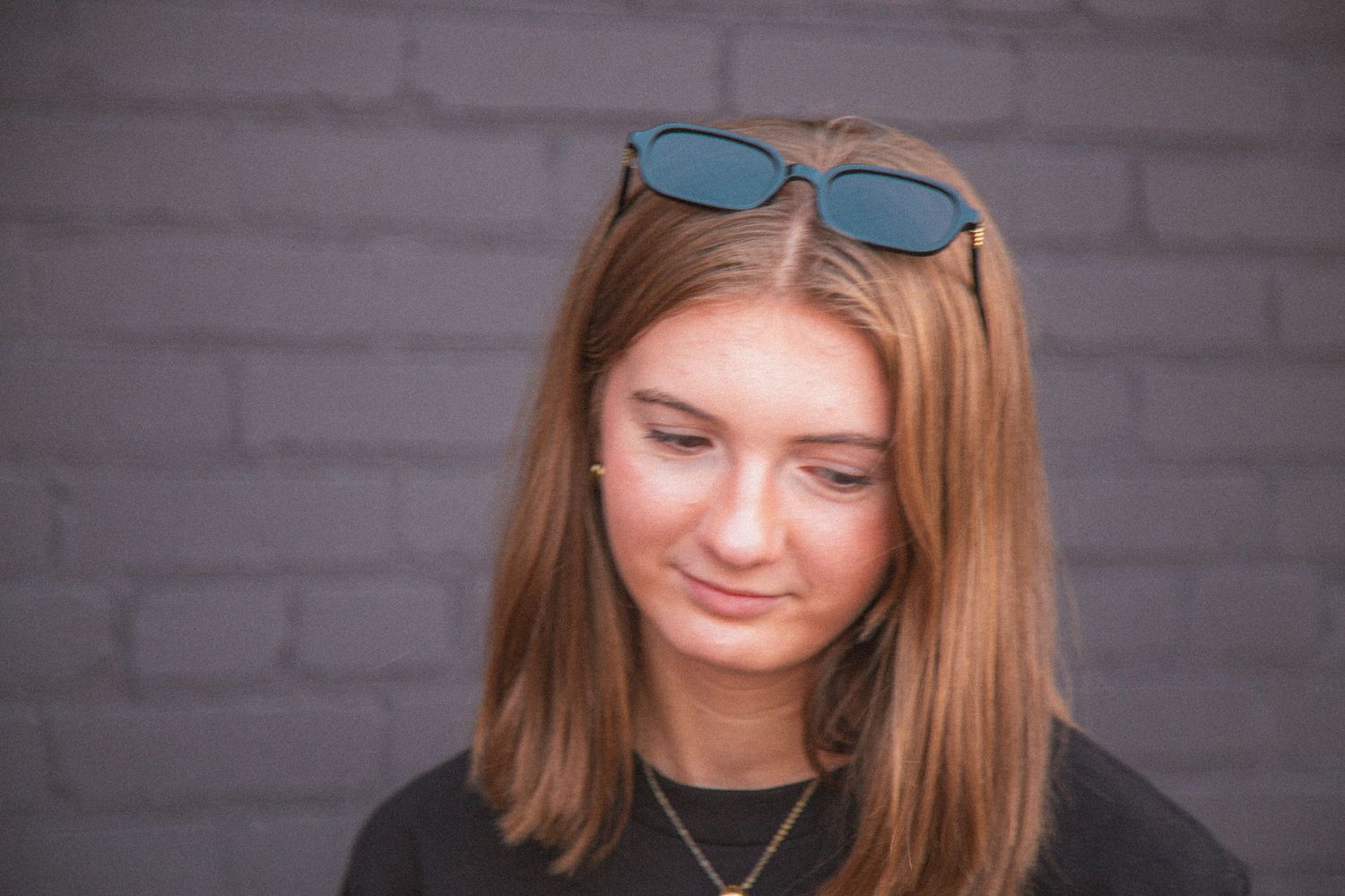 Young woman with sunglasses on head looking down, wearing black tee against gray brick wall.