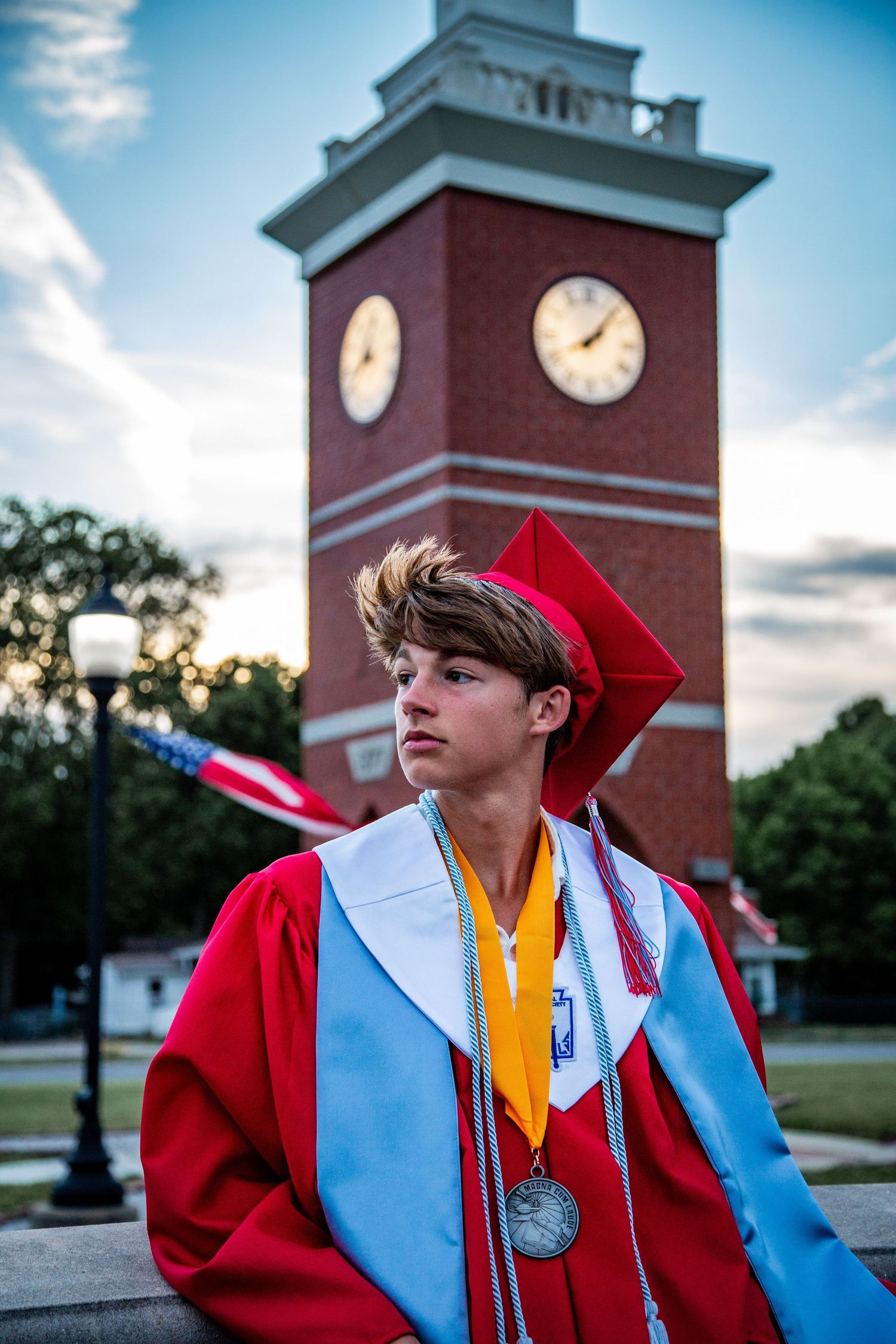 High school graduate in red cap and gown posing thoughtfully in front of a brick clock tower at dusk.